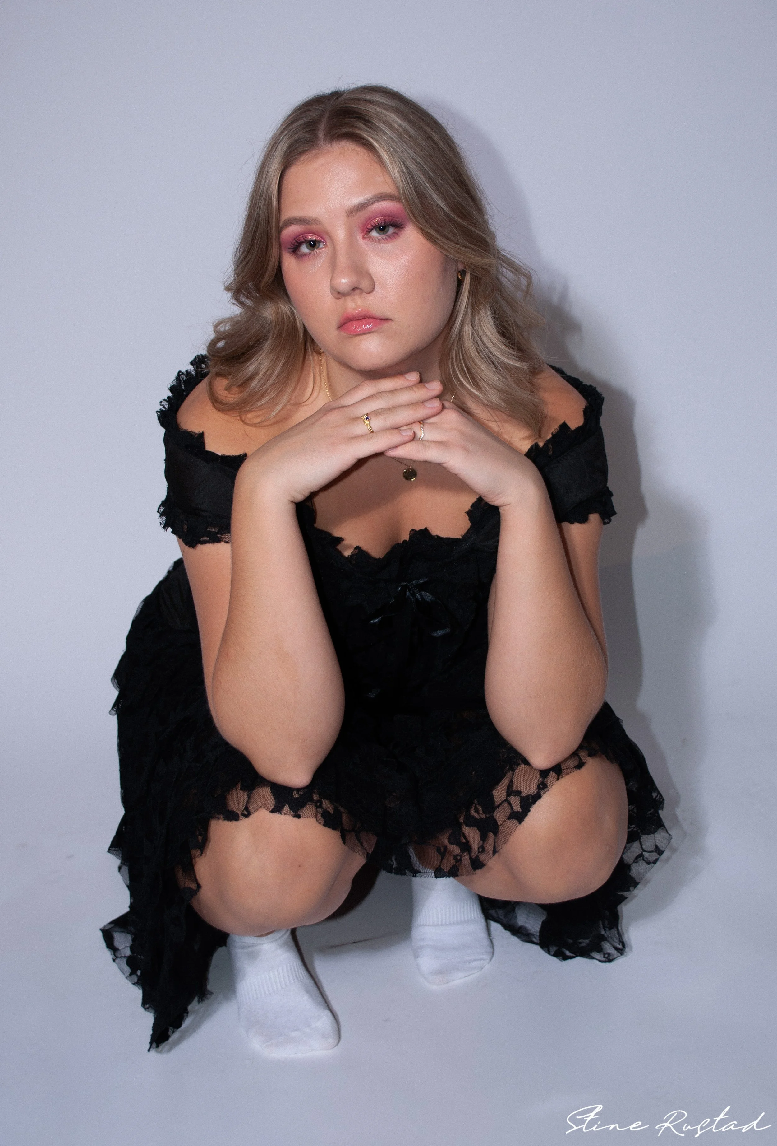 A young woman with blonde hair and pink makeup squats against a plain gray background, wearing a black lace dress, white socks, and jewelry.