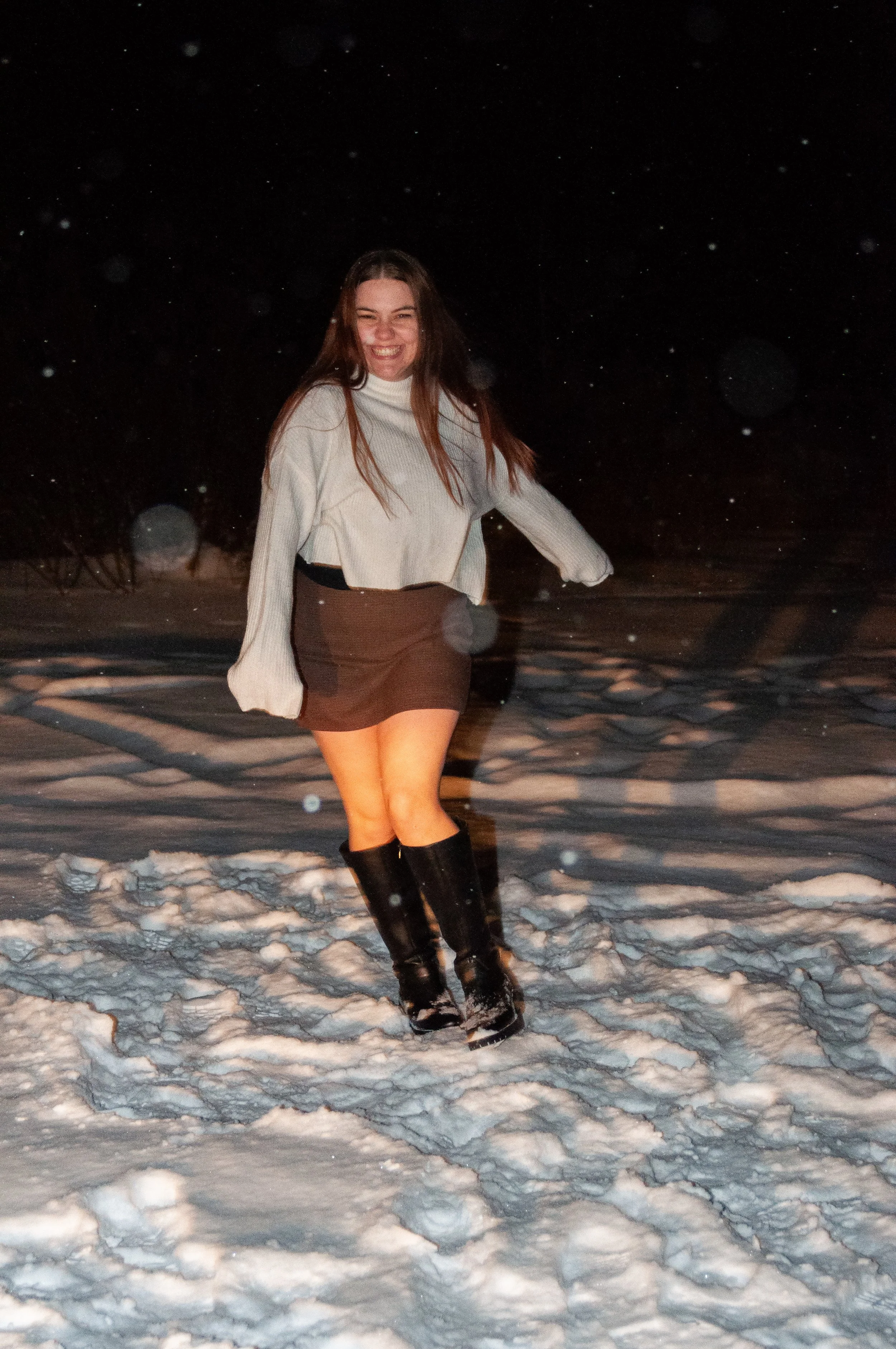 Young woman smiling and playing in snow at night, wearing a white sweater, dark skirt, and black boots.