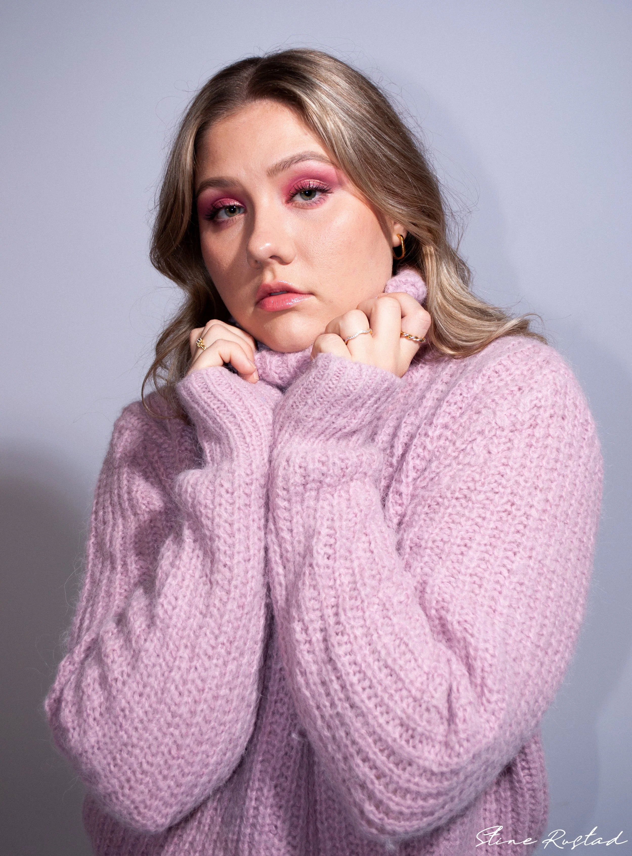 A young woman with blonde hair and pink makeup, wearing a pink knitted sweater, posing with her hands near her face against a neutral background.