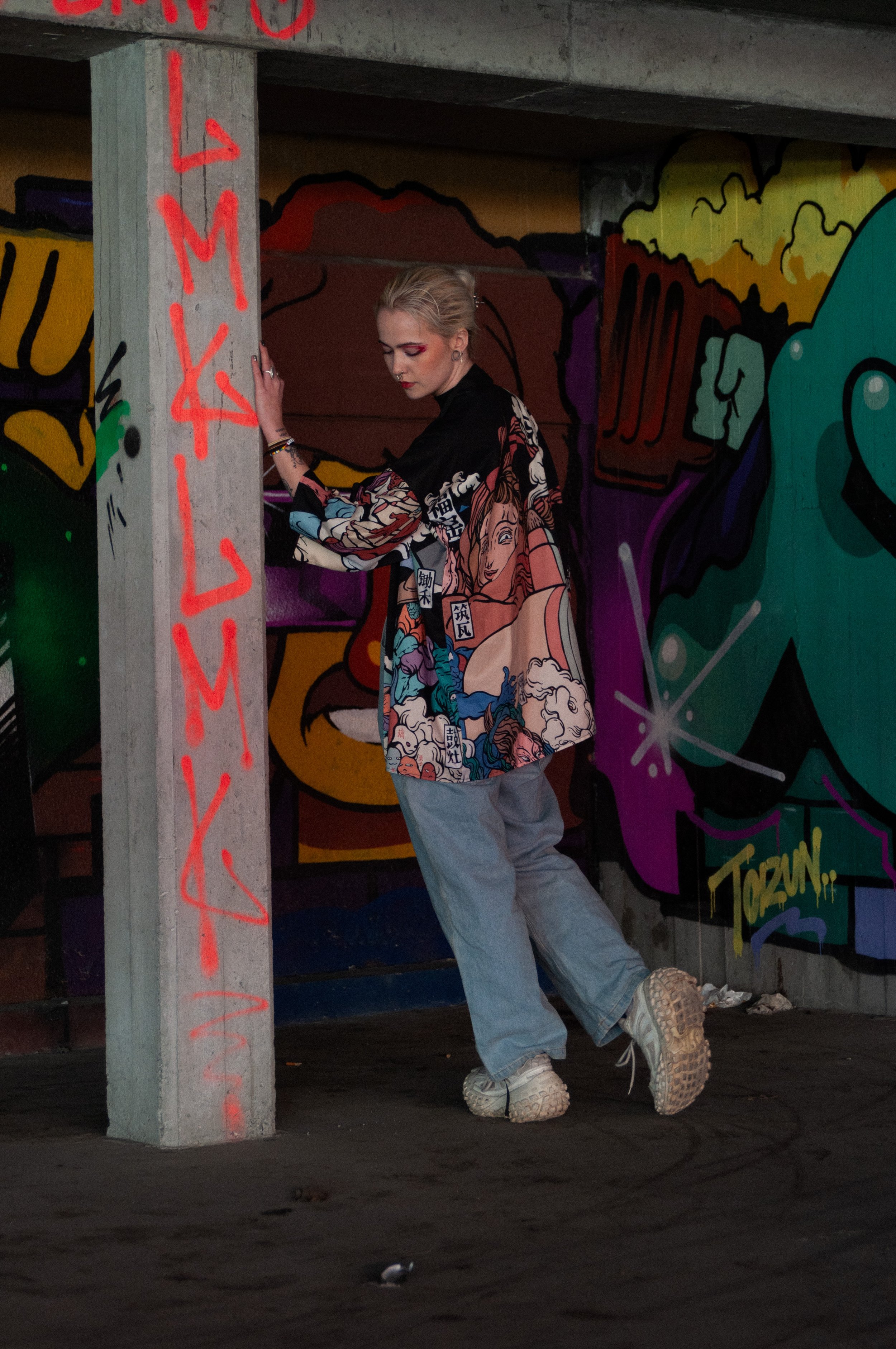 A young woman with blonde hair in a bun, wearing a colorful graphic oversize shirt, light gray pants, and white sneakers, stands under a graffiti-covered concrete structure, touching a concrete pillar with red graffiti text, in front of a vibrantly p