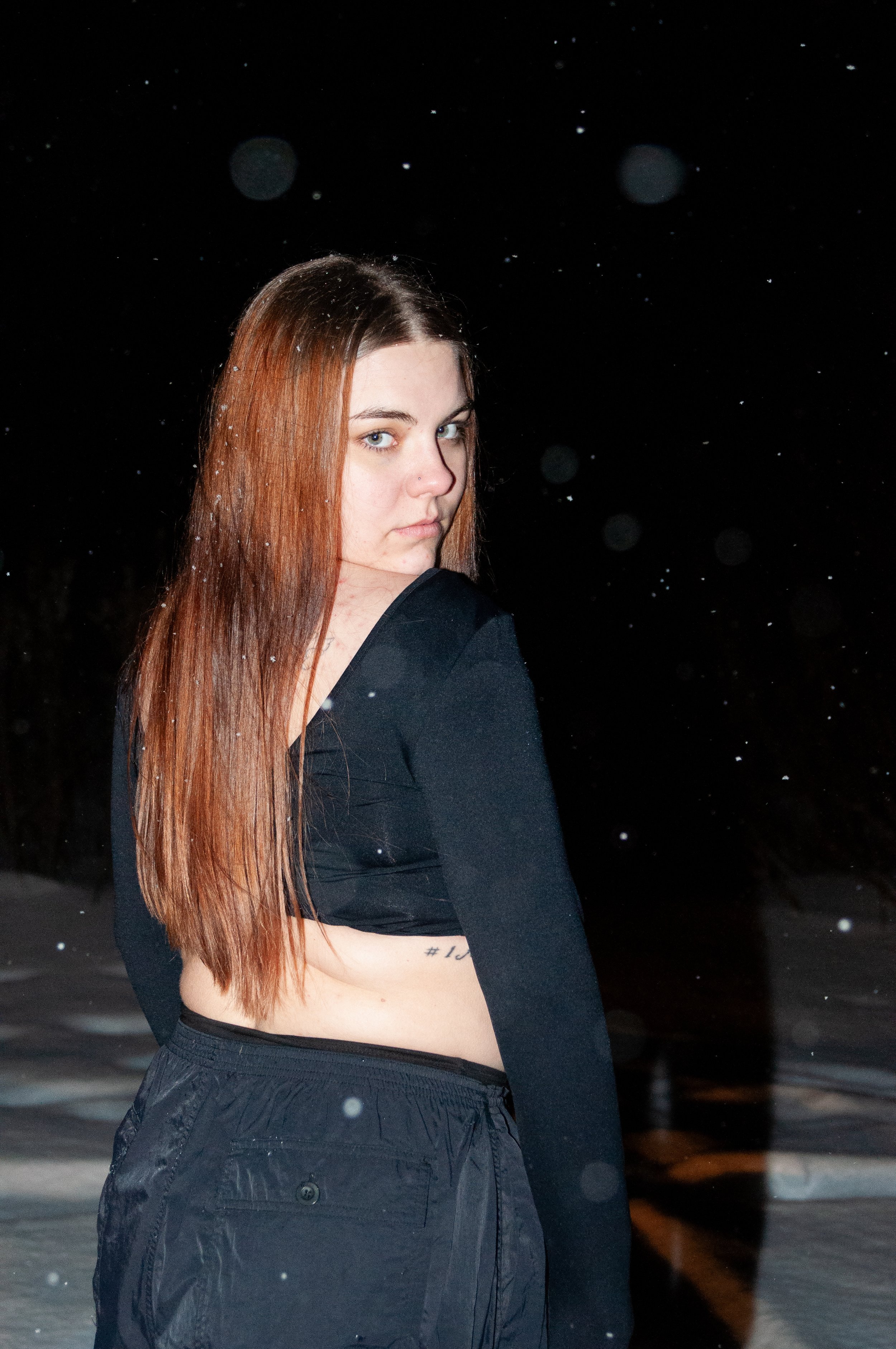 A young woman with long, reddish-brown hair looks over her shoulder at night, with snow falling around her and a dark background.