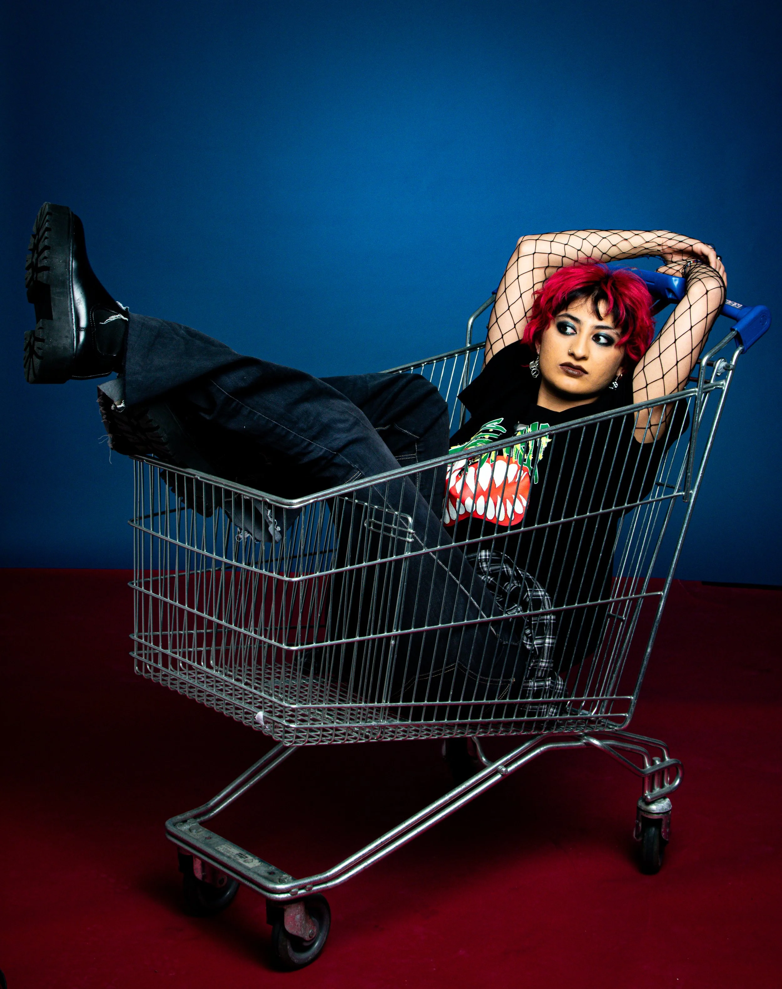 A young person with vibrant pink hair and edgy clothing reclines inside a shopping cart against a blue background.