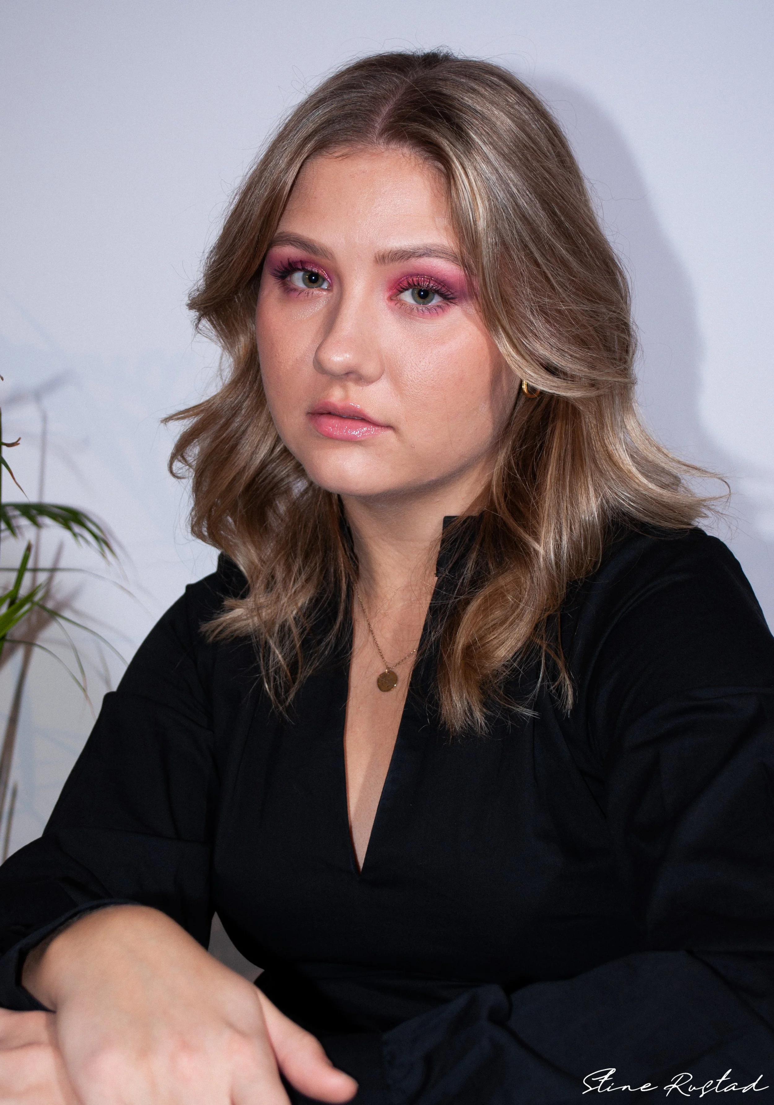 Portrait of a woman with shoulder-length wavy light brown hair, wearing pink eyeshadow, a black top, a gold necklace, and small hoop earrings, sitting in front of a white background with a green plant in the corner.
