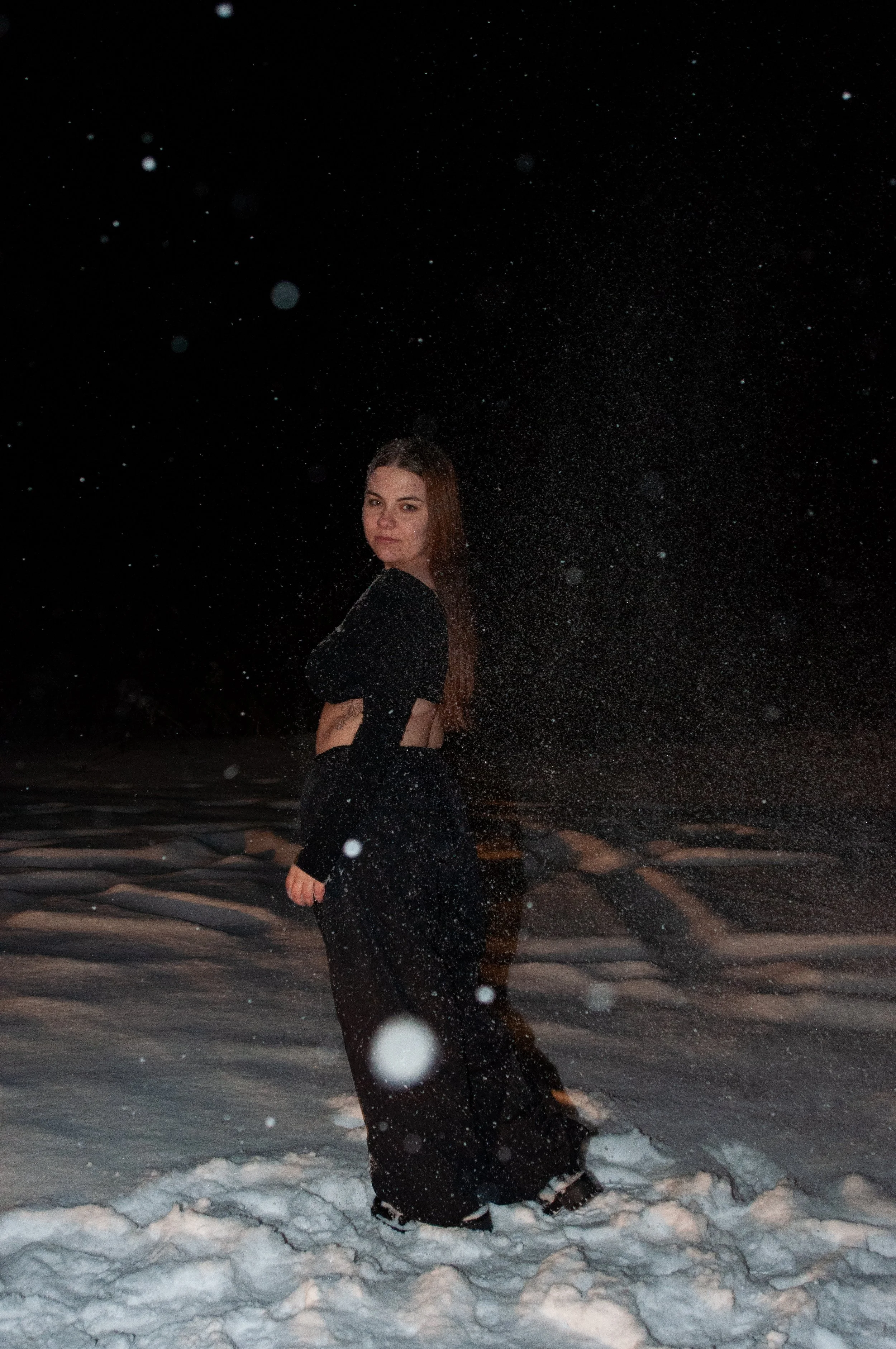 A woman standing on snow at night with snowflakes falling around her, dressed in black, looking at the camera.