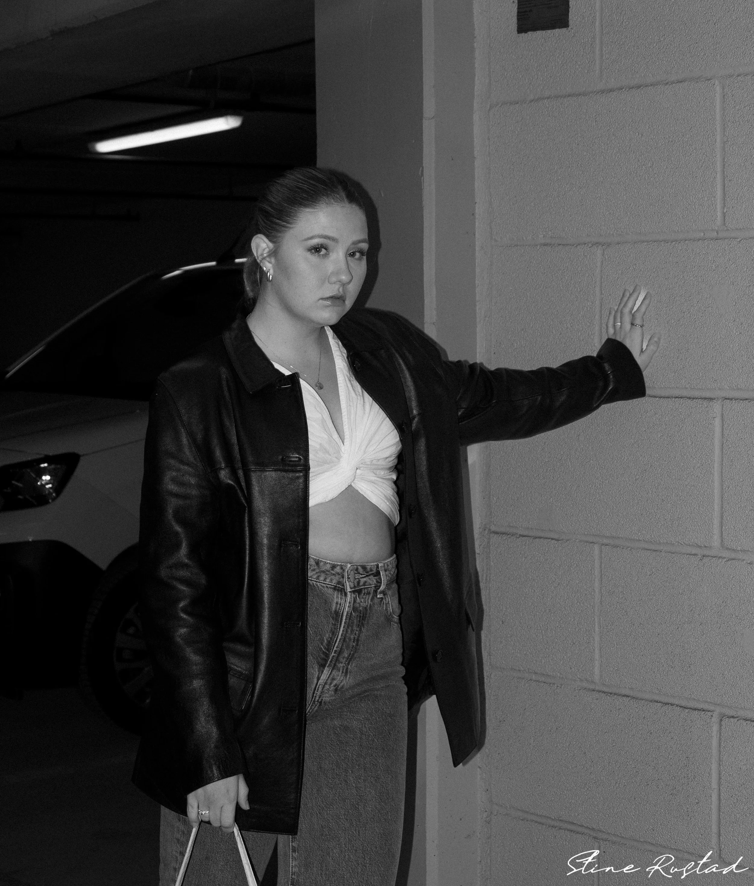 A young woman with light skin, dark hair in a ponytail, wearing a black leather jacket, a knotted white crop top, and high-waisted jeans, standing in a parking garage with a concrete wall behind her. She is leaning against the wall with her right han