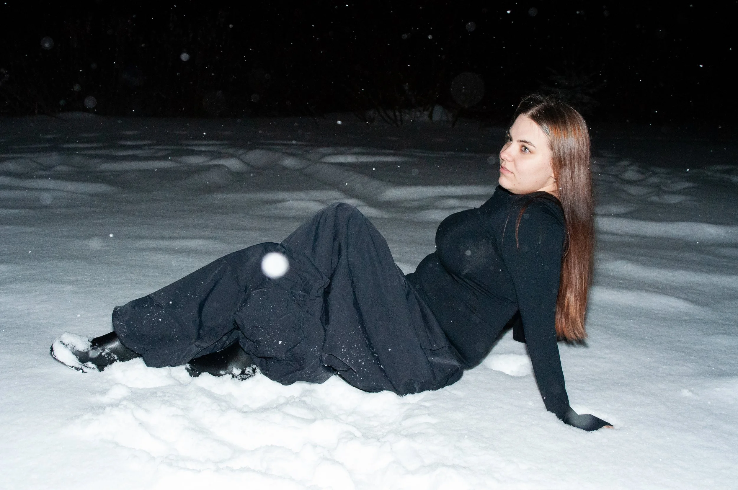 A woman in black winter clothing sitting on snow at night with falling snowflakes.