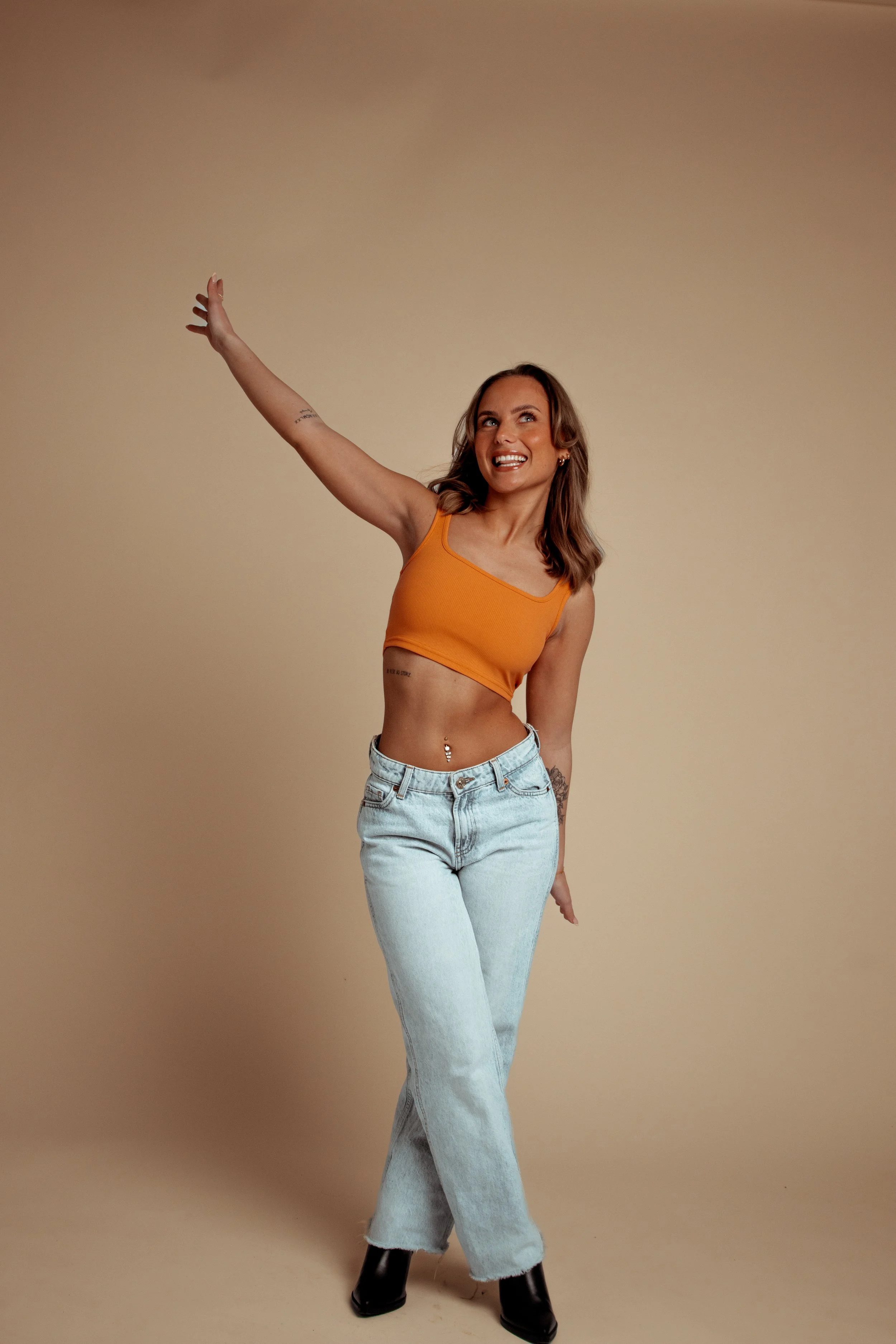 A woman with shoulder-length brown hair wearing an orange crop top, light blue jeans, and black boots, smiling and raising her right arm against a beige background.