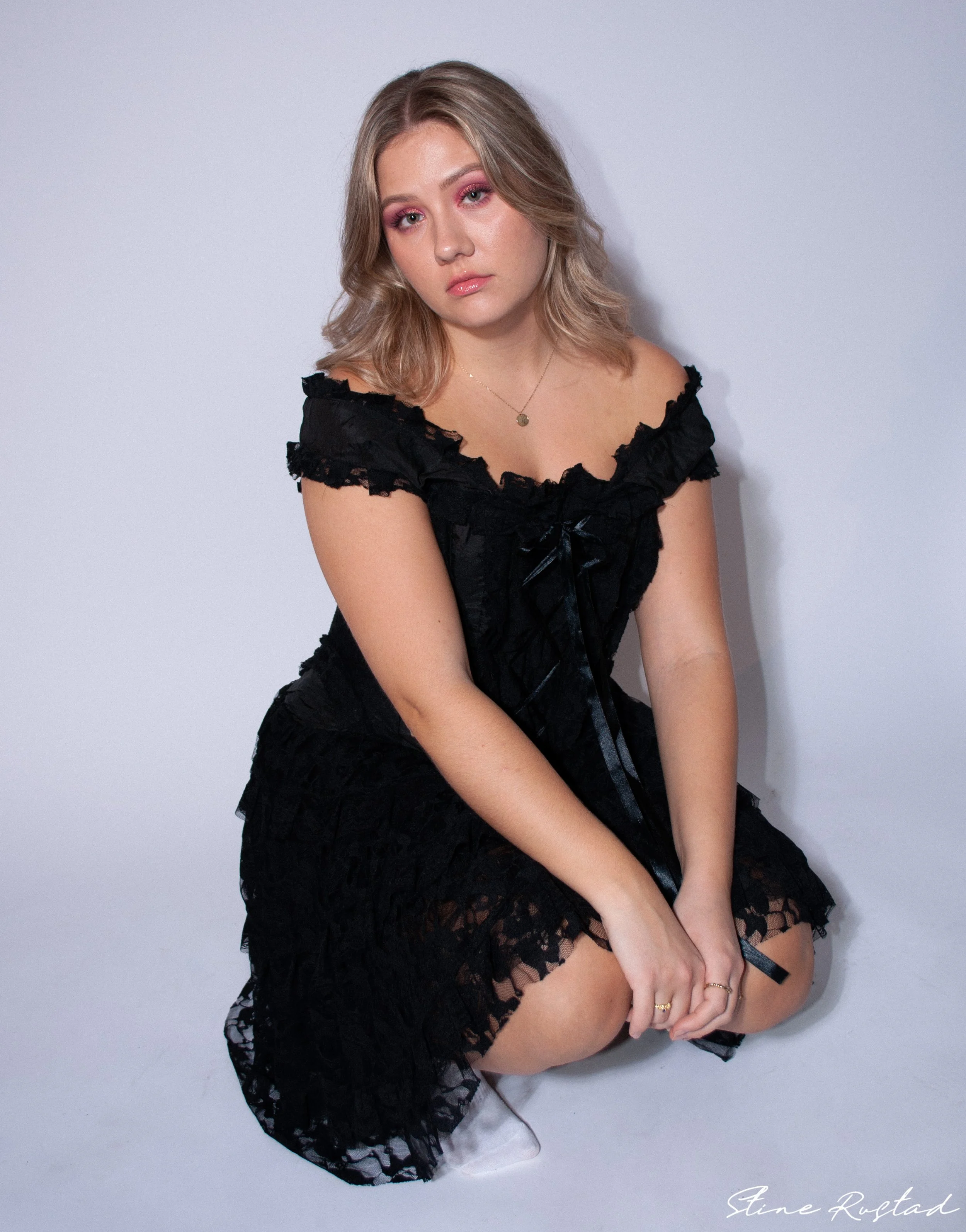 Young woman with blonde hair and pink makeup, wearing a black lace dress, sitting on the floor against a plain white background.