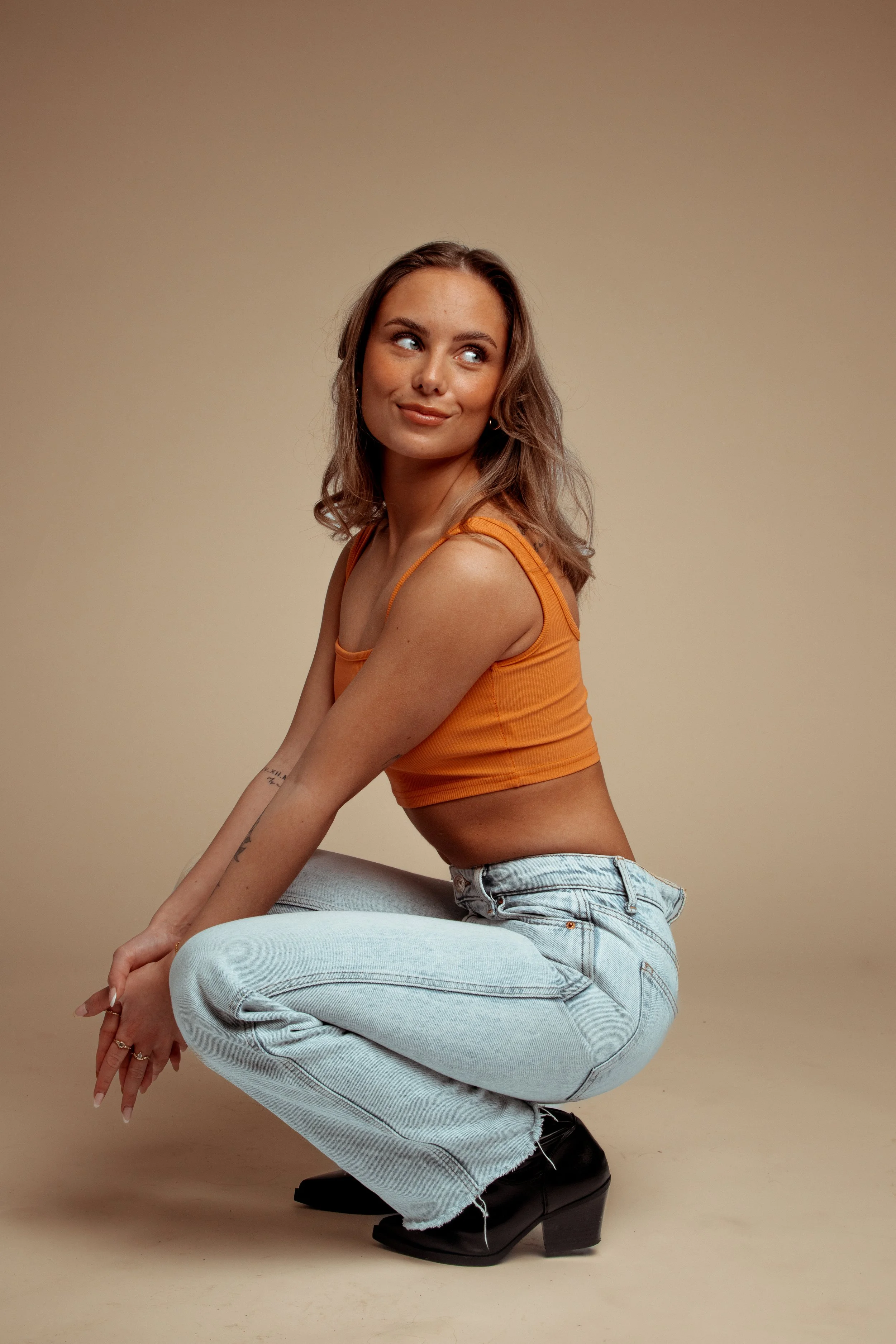 A young woman with wavy hair wearing an orange crop top, high-waisted jeans, and black platform shoes, squatting against a beige background, looking to her left.