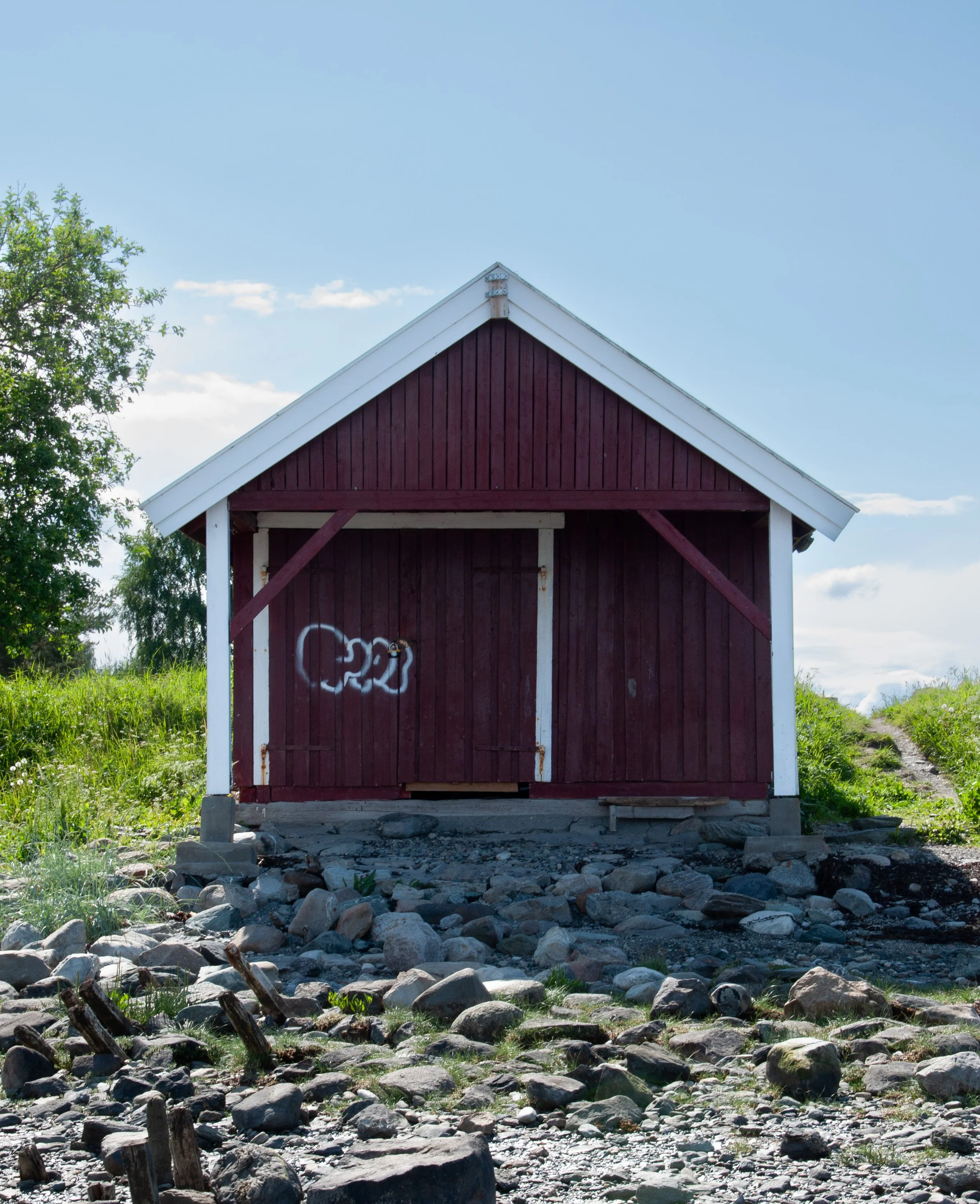 A wooden red and white shed or small barn on rocky ground, with graffiti on the door and a gravel pathway on the right, under a blue sky with clouds and surrounded by green grass and trees.