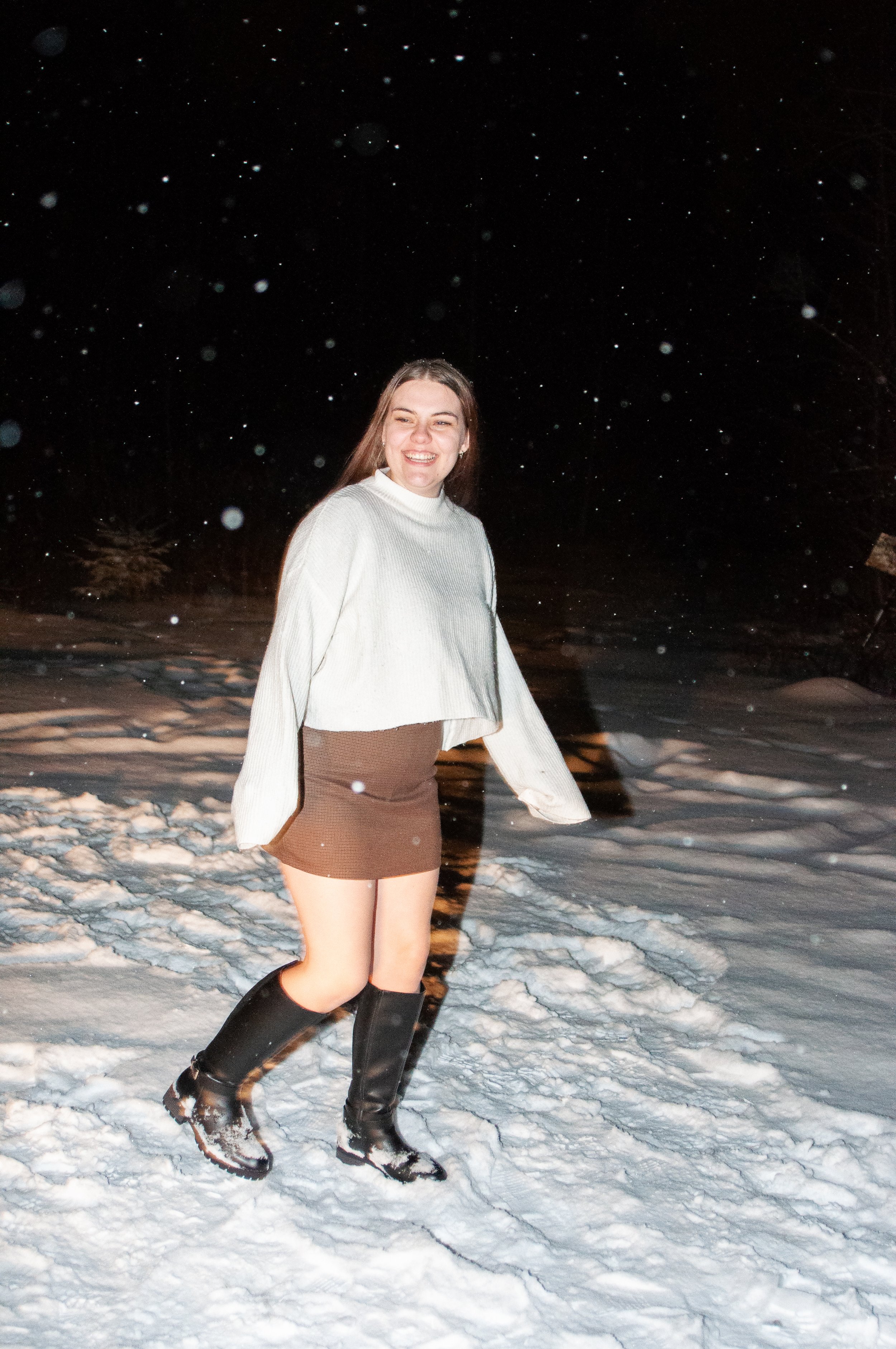A young woman with long brown hair smiling while standing in snow at night, wearing a white long-sleeve sweater, a brown skirt, and black rain boots, with snow falling around her.