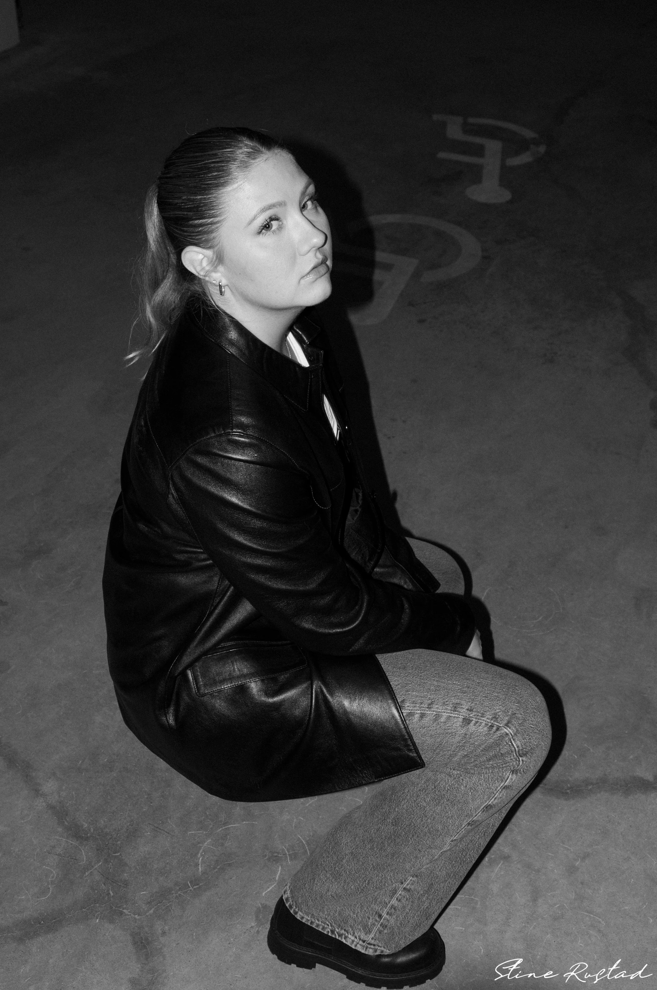 A woman in a leather jacket and jeans is crouching on a concrete floor, looking up at the camera with a serious expression. There is a faded number or symbol on the ground nearby.