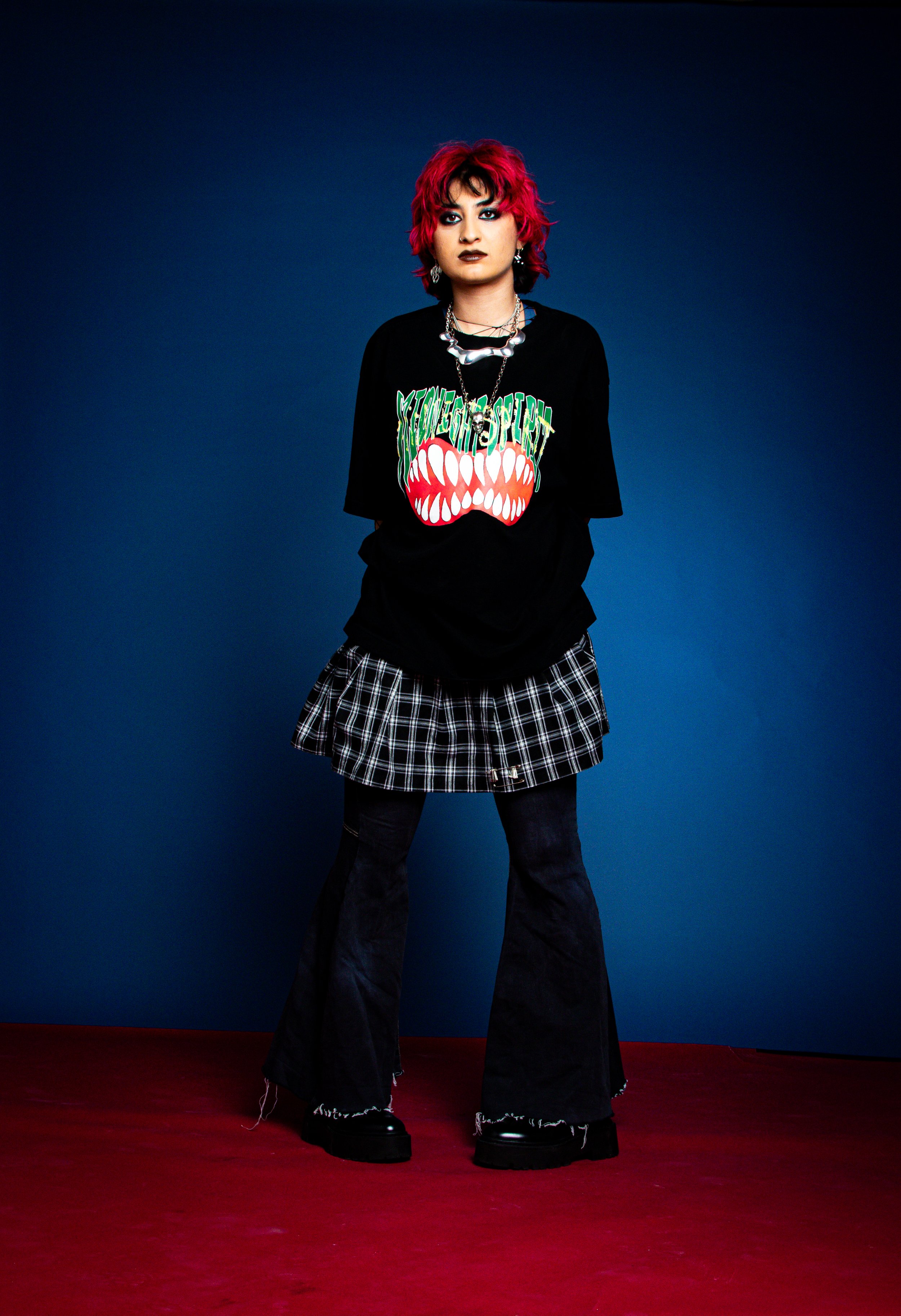A person with short, curly pink hair and dark makeup stands against a dark blue background. They are wearing a black graphic t-shirt, a plaid skirt layered over black pants, chunky black shoes, and accessorized with jewelry.