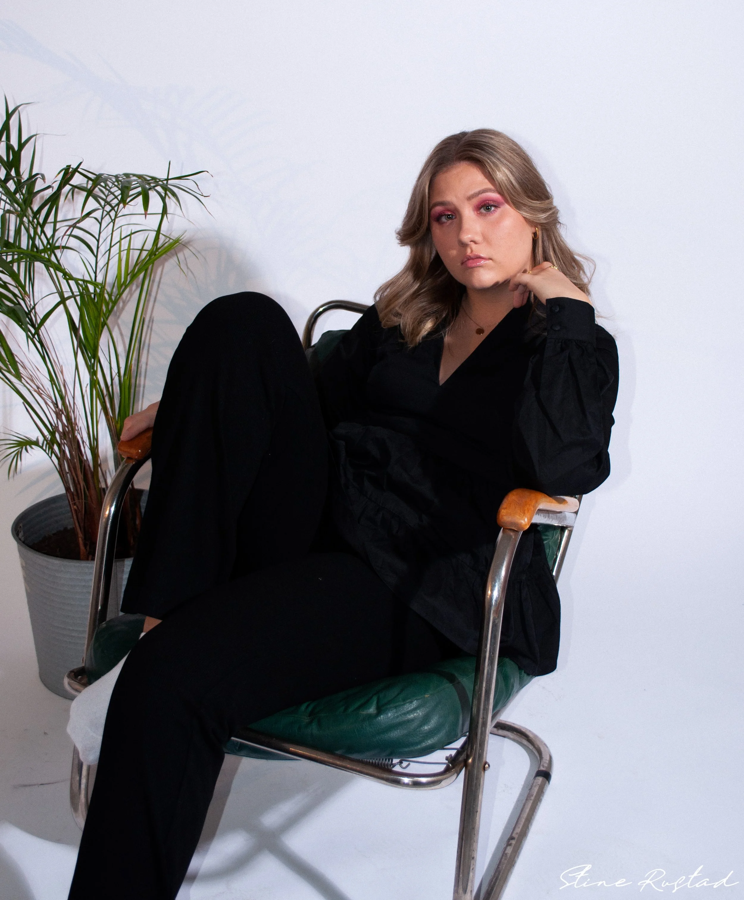A young woman with blonde wavy hair sits on a vintage green leather and metal chair against a plain white background. She is wearing a black long-sleeve top and black pants, with pink eyeshadow and gold jewelry. There is a potted plant with long, thi