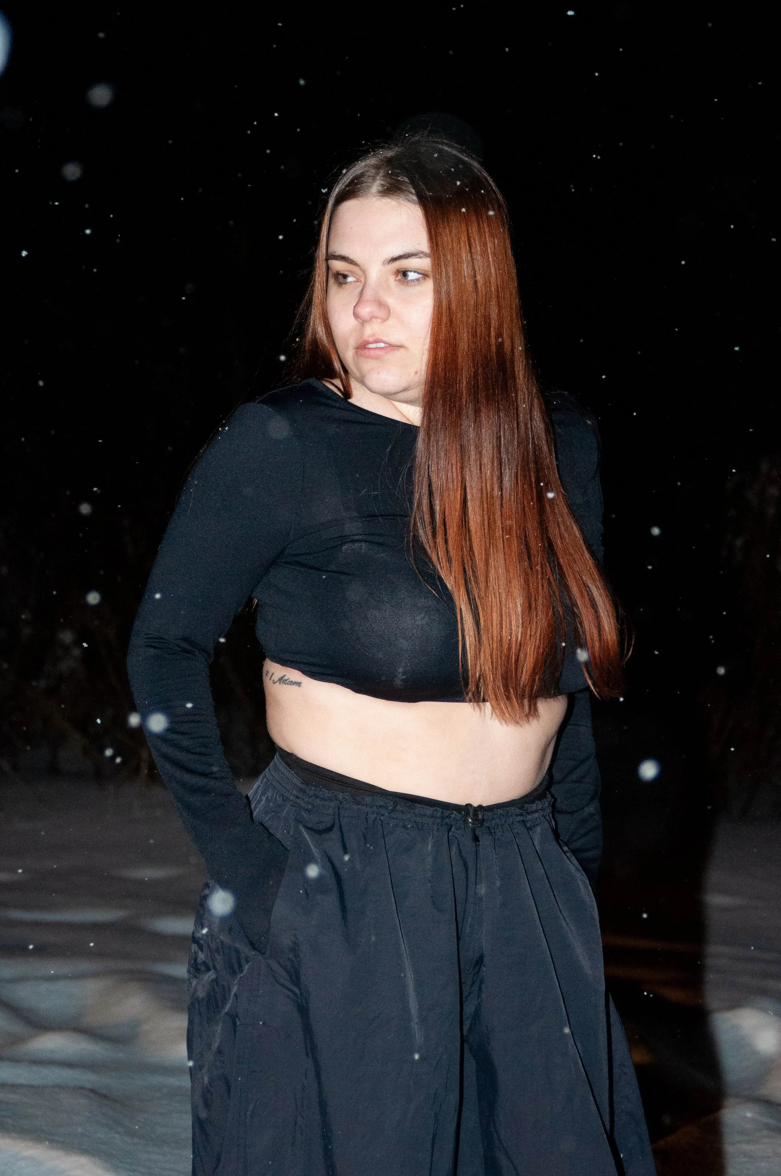 A young woman with long red hair standing outdoors at night in the snow, wearing a black crop top and black pants.