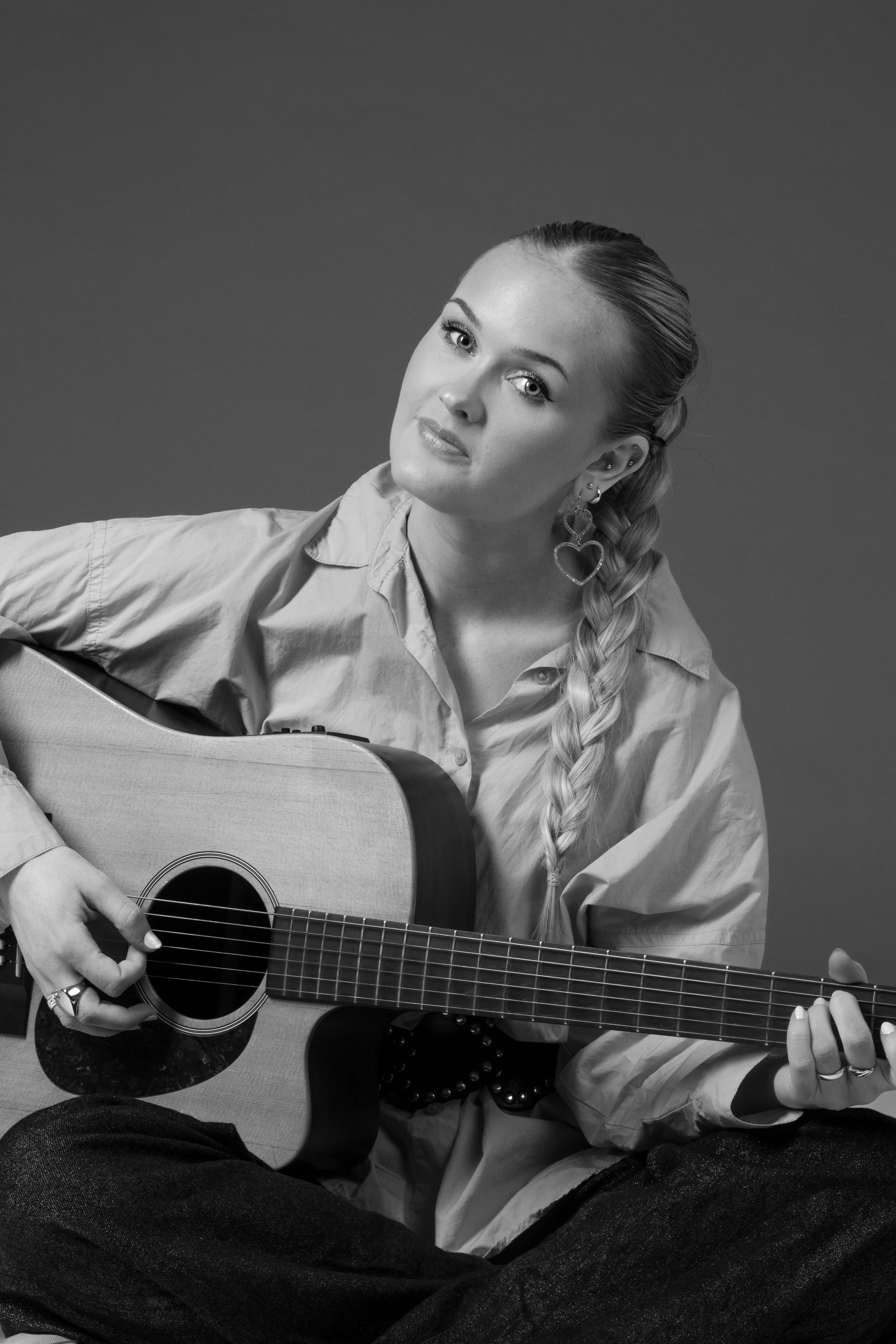 Black and white portrait of a young woman with braided hair, wearing earrings and a loose shirt, sitting cross-legged while playing an acoustic guitar.