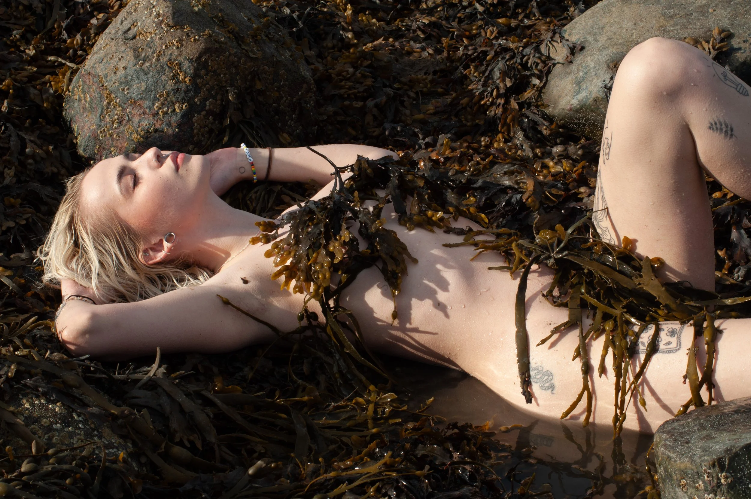 A woman with blonde hair relaxing on a bed of seaweed, surrounded by rocks, with her eyes closed and one arm behind her head.