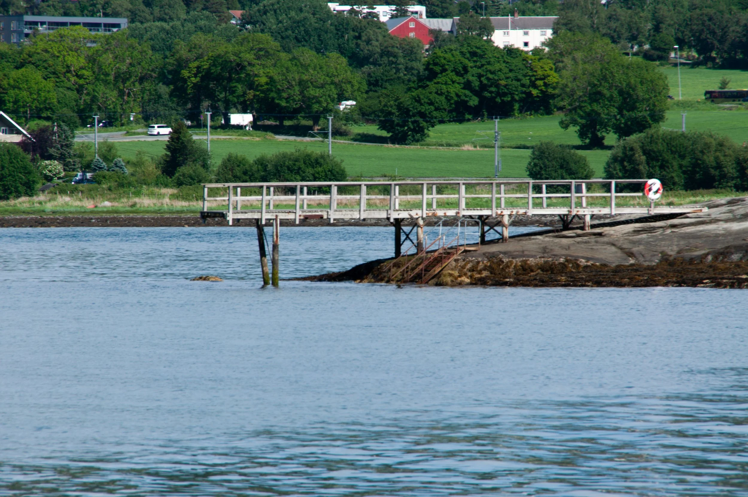 A wooden pier extending over water with a life buoy attached at the end, over a rocky shoreline with greenery and trees in the background.