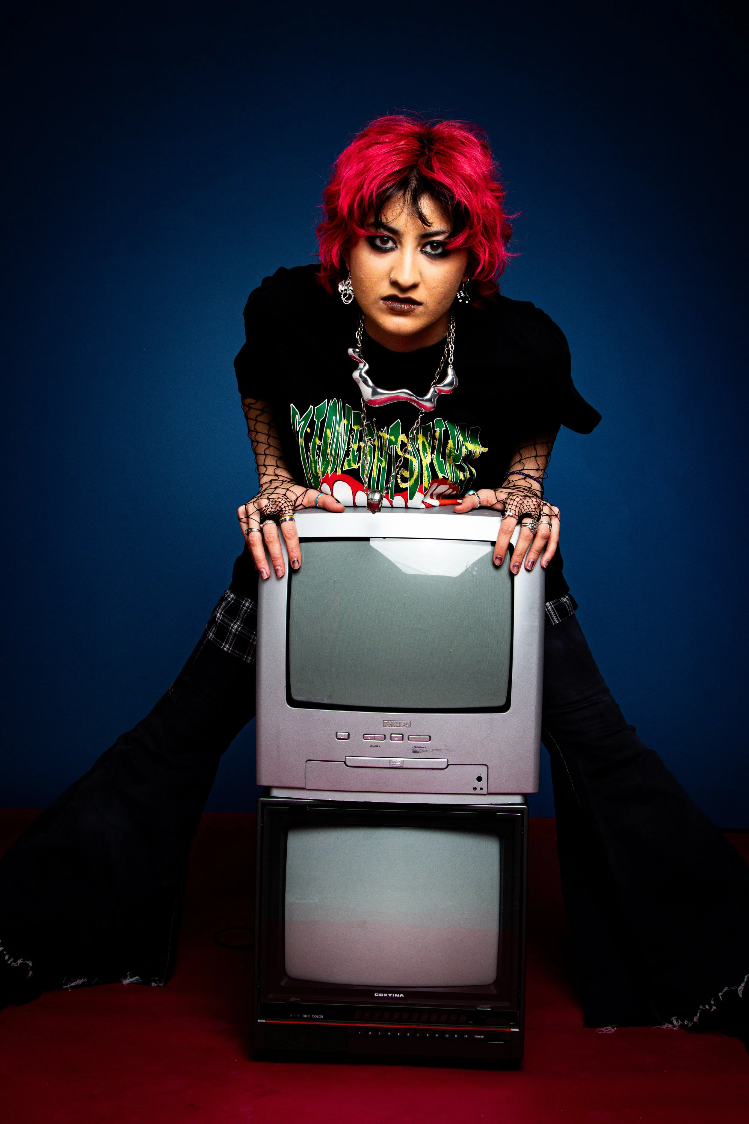 A woman with vibrant pink hair, dark eye makeup, and gothic style clothing leans over a stack of two old televisions, set against a dark background.