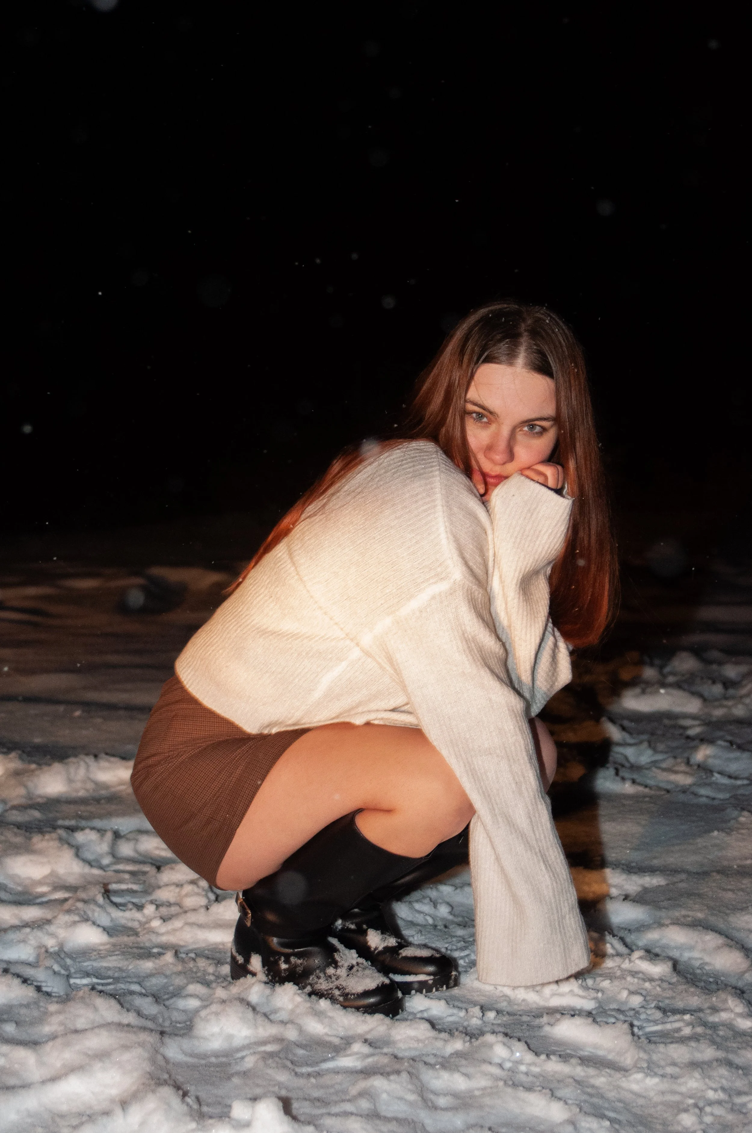 A woman crouching in the snow at night, wearing a cream sweater, brown skirt, and black boots, with snow falling around her.