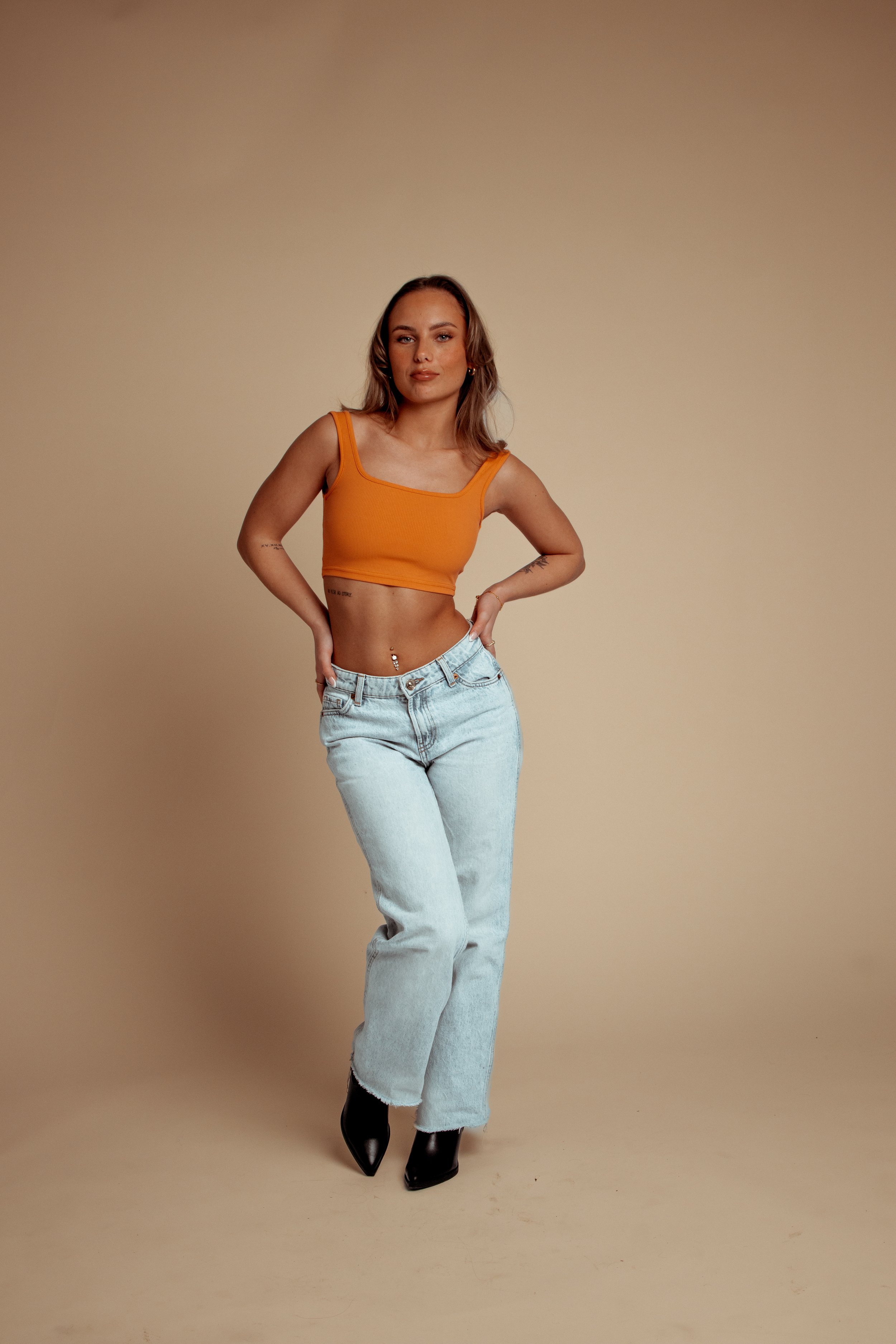 A woman standing in front of a beige background, wearing an orange crop top, light wash jeans, and black high boots.