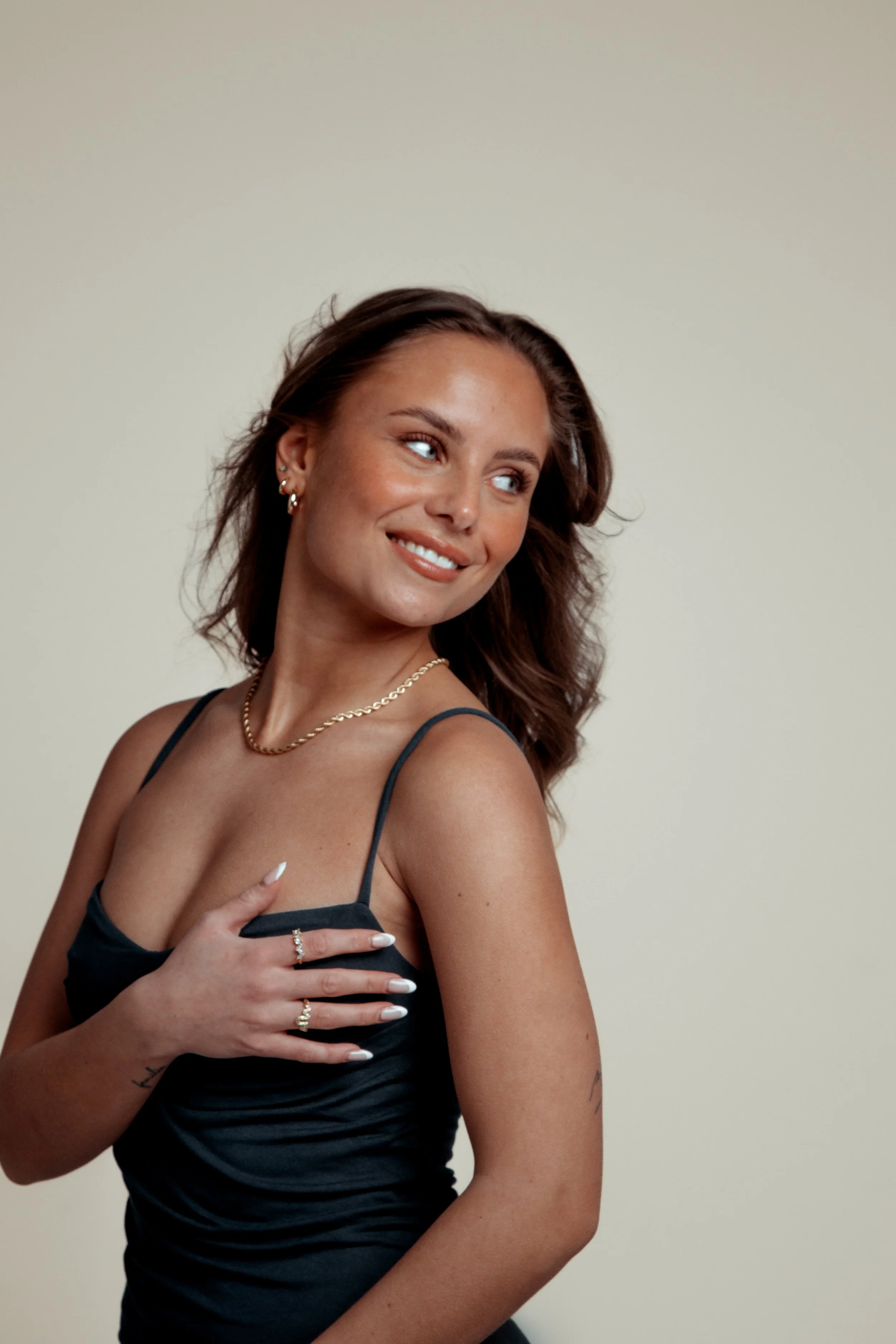 A woman with shoulder-length brown hair, wearing a black spaghetti strap dress, jewelry, and with her right hand over her chest, smiling and looking to her left.