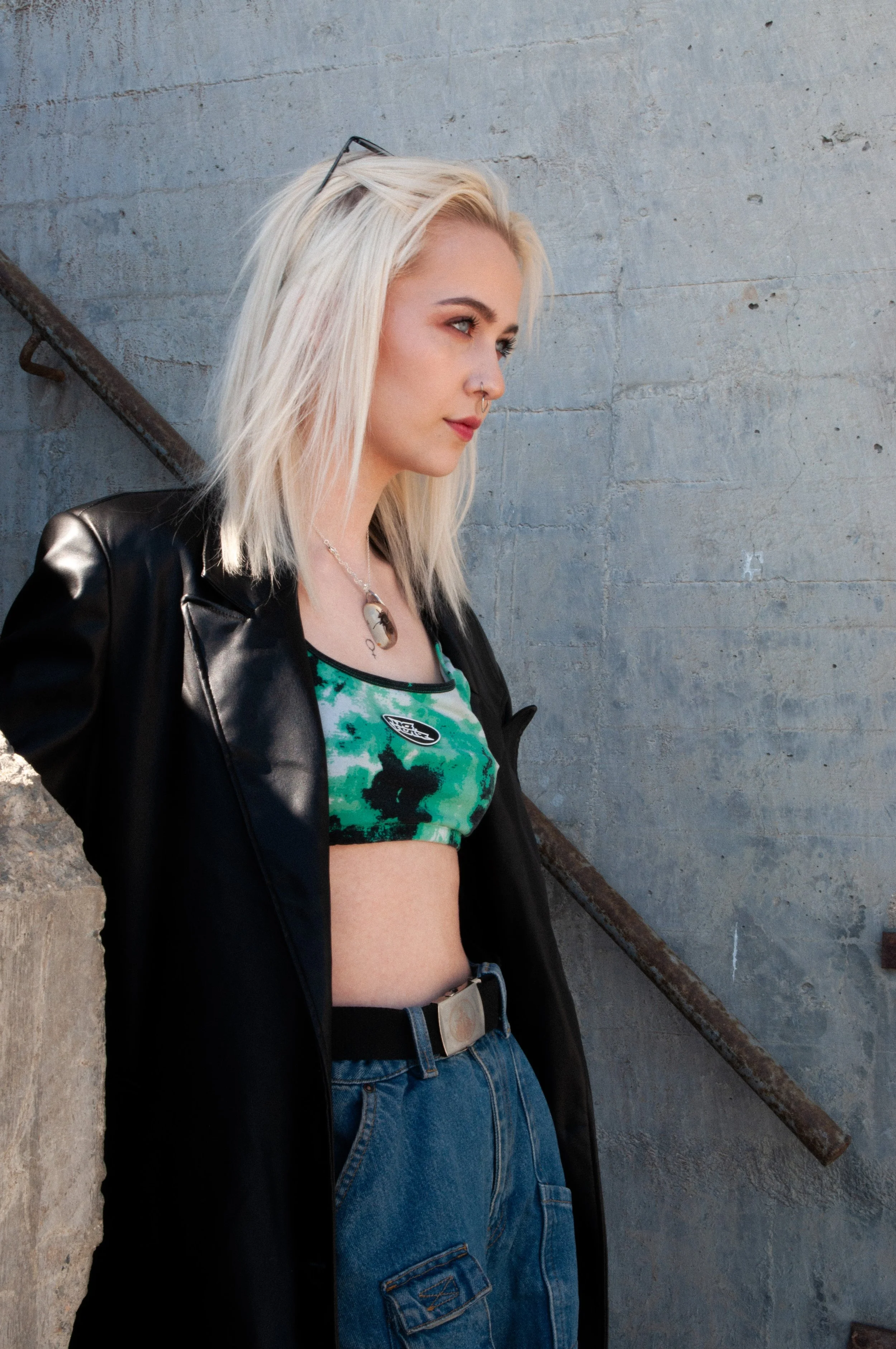 Young woman with blonde hair and multiple piercings, wearing a black leather jacket and green tie-dye crop top, standing against a textured gray concrete wall with a rusty metal pipe.