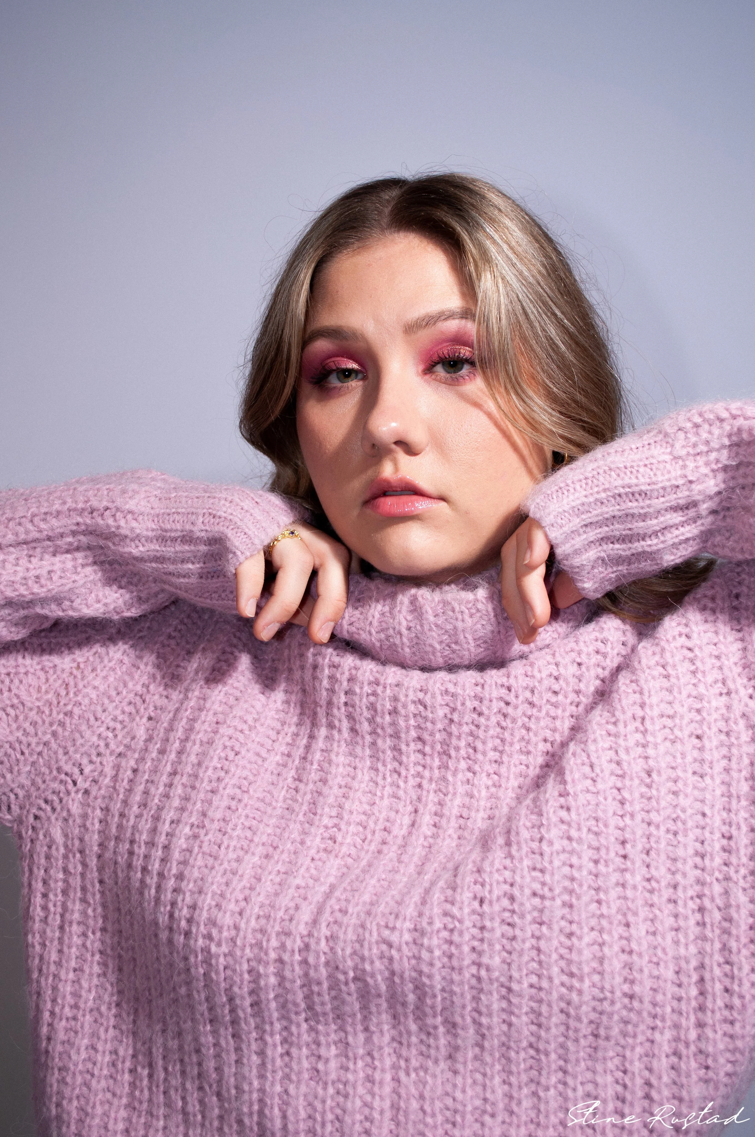 A woman with pink eyeshadow wearing a pink knit sweater rests her chin on the collar of her sweater, looking at the camera with a neutral expression.