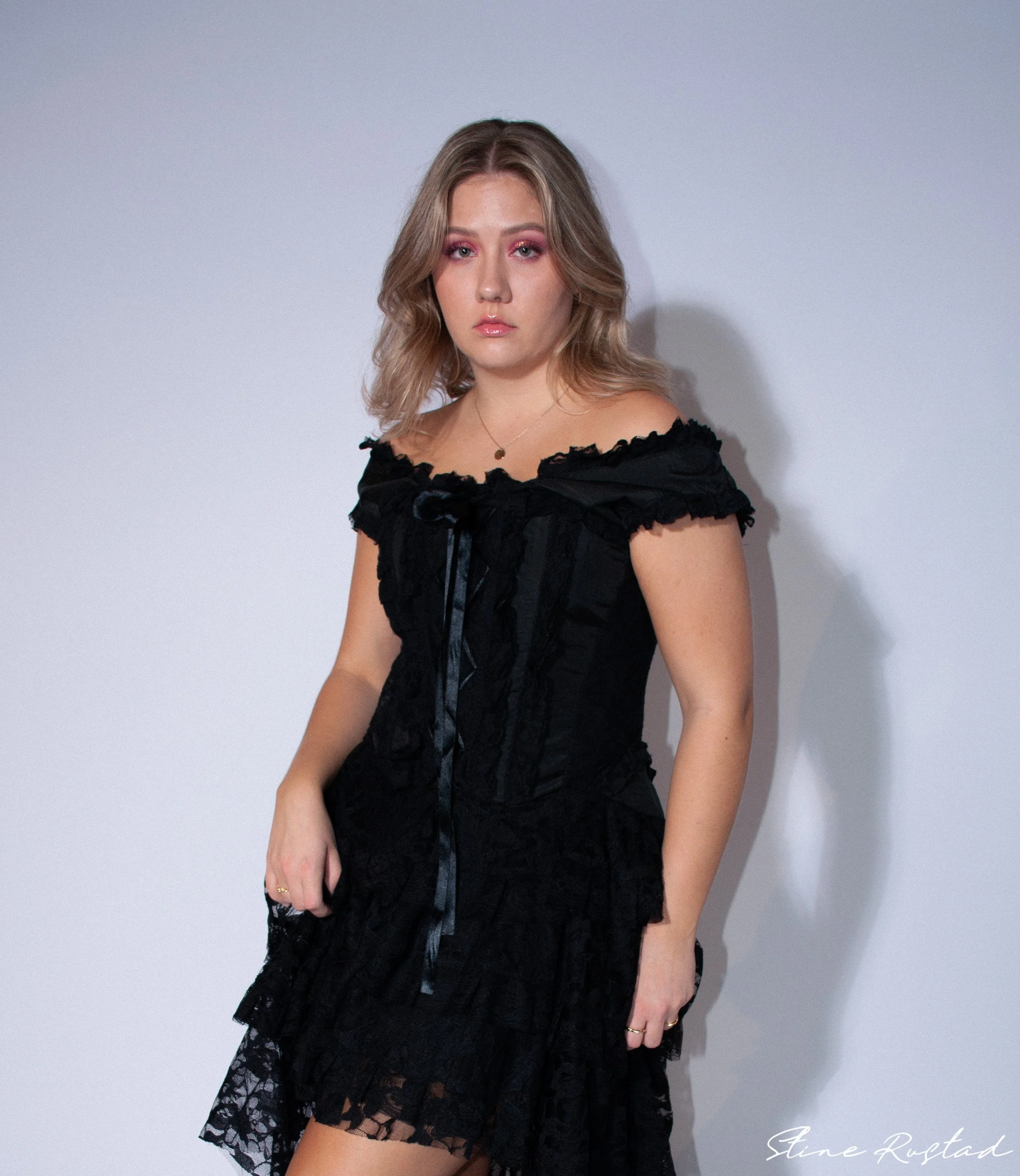 A young woman with blonde hair and pink eyeshadow wearing a black off-the-shoulder dress with lace details, standing against a plain light-colored wall.