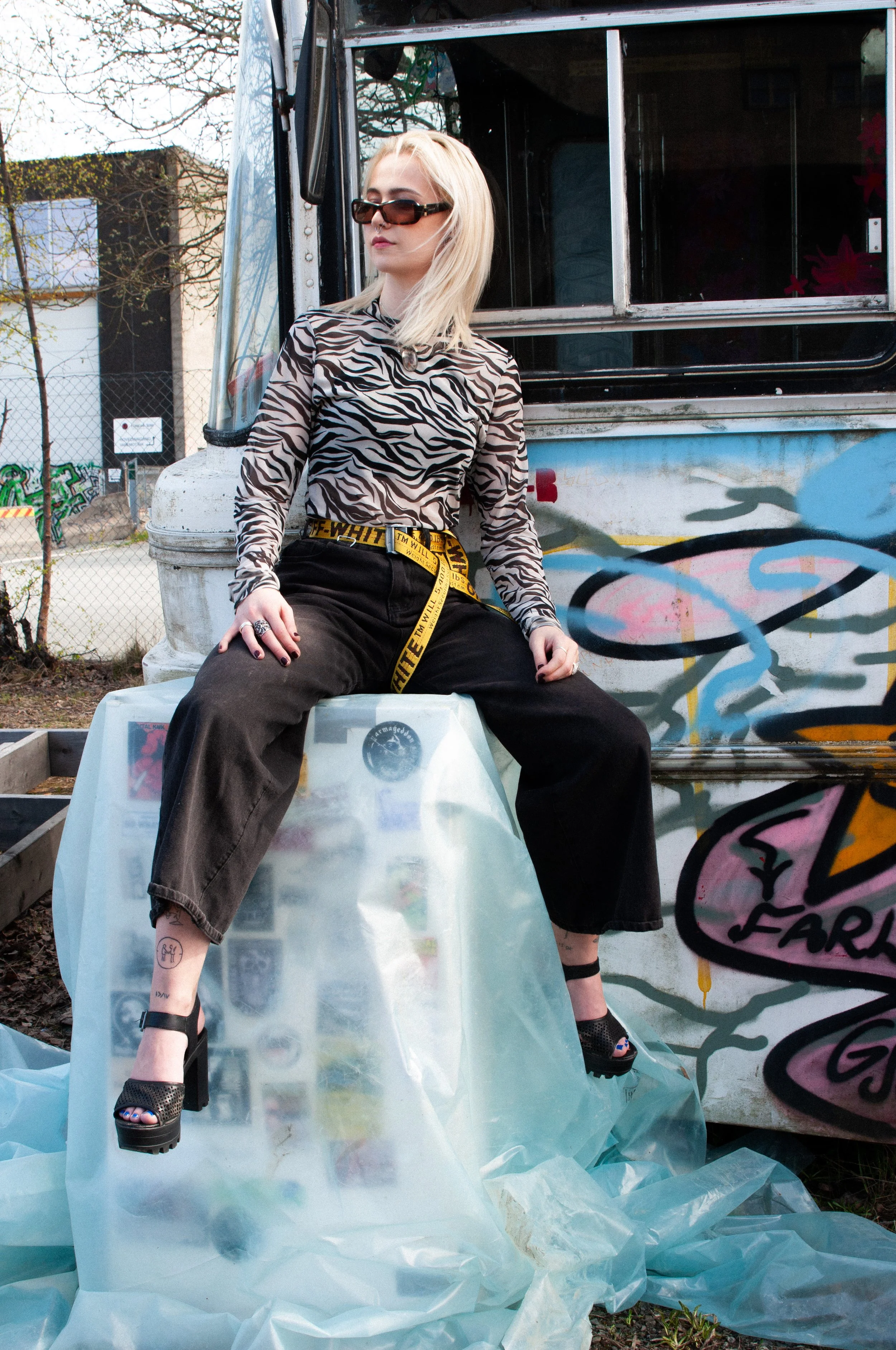 A woman with blonde hair in sunglasses, wearing a zebra print top, black pants, and high heels, sitting on a plastic-covered surface in front of a graffiti-covered food truck.