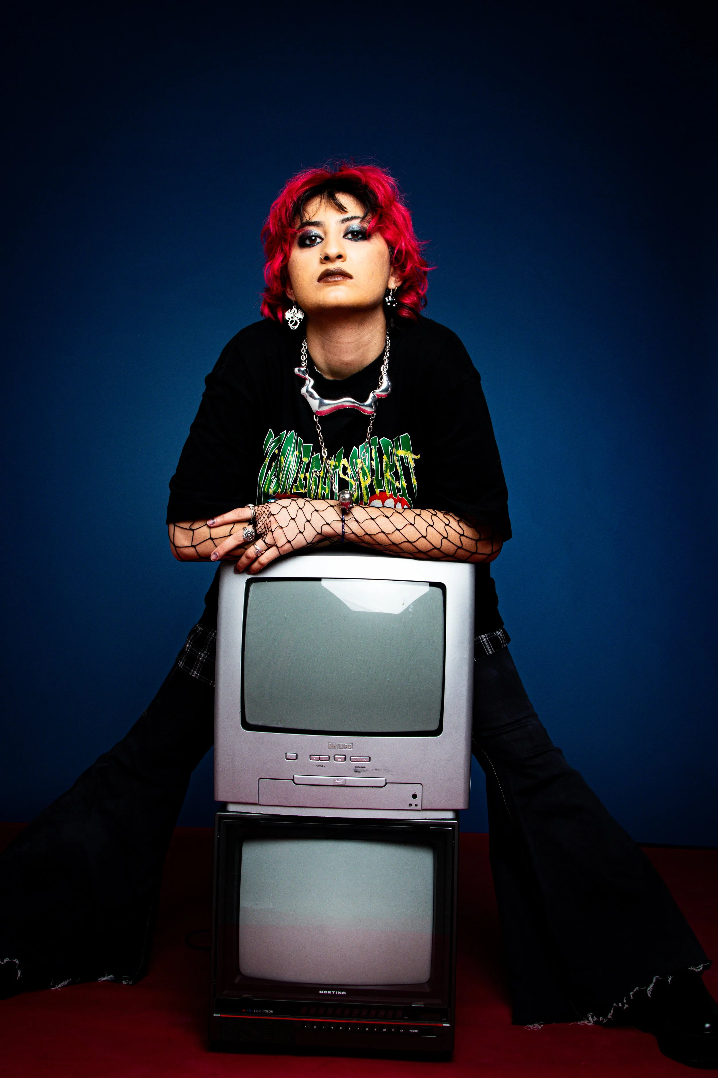 A person with bright pink hair leaning on two stacked vintage televisions against a dark blue background.