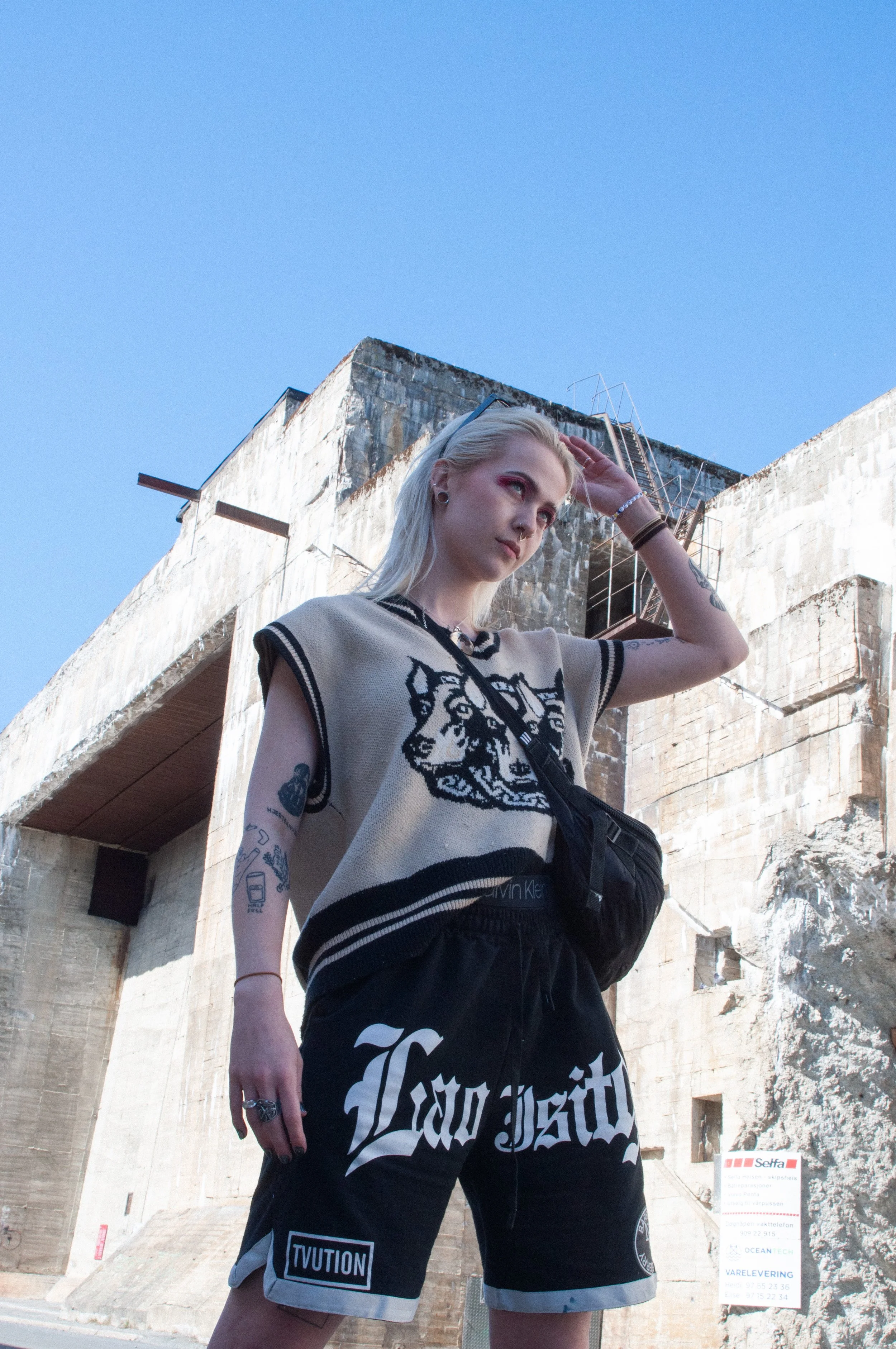 Young woman with tattoos, blonde hair, and casual streetwear pose outdoors against a large, weathered concrete building with stairs and clear blue sky.