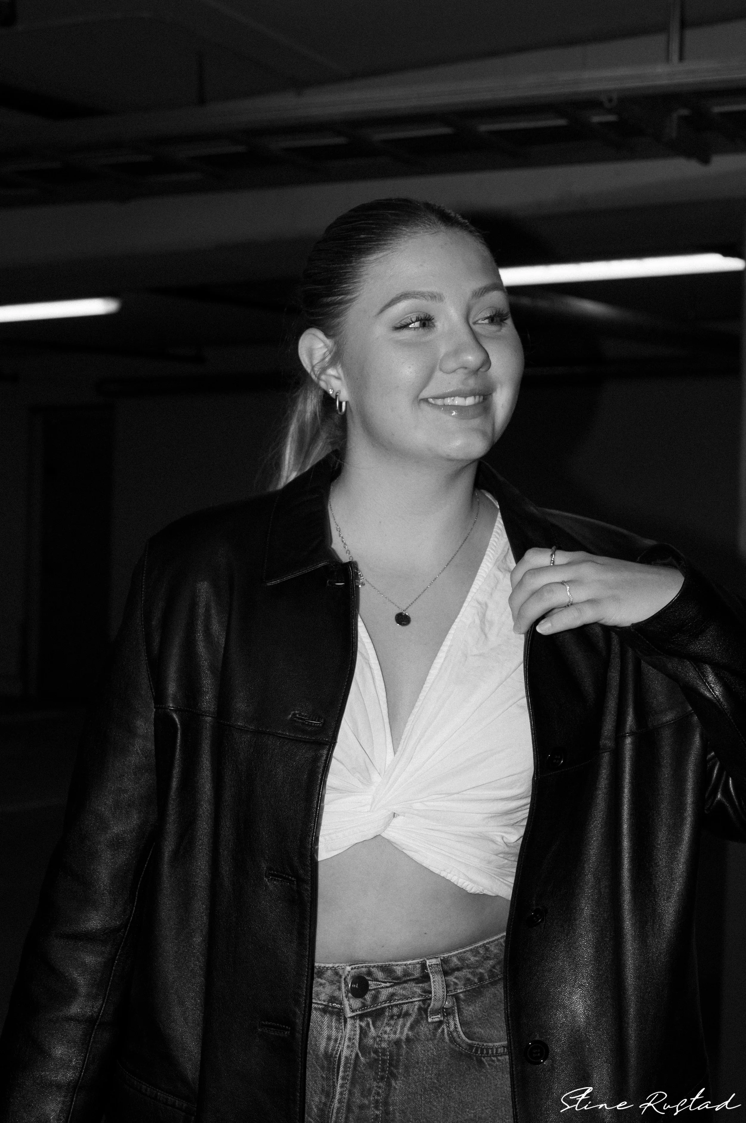 A young woman smiling with her eyes closed, wearing a leather jacket, a knotted white shirt, earrings, necklaces, and rings, standing indoors.
