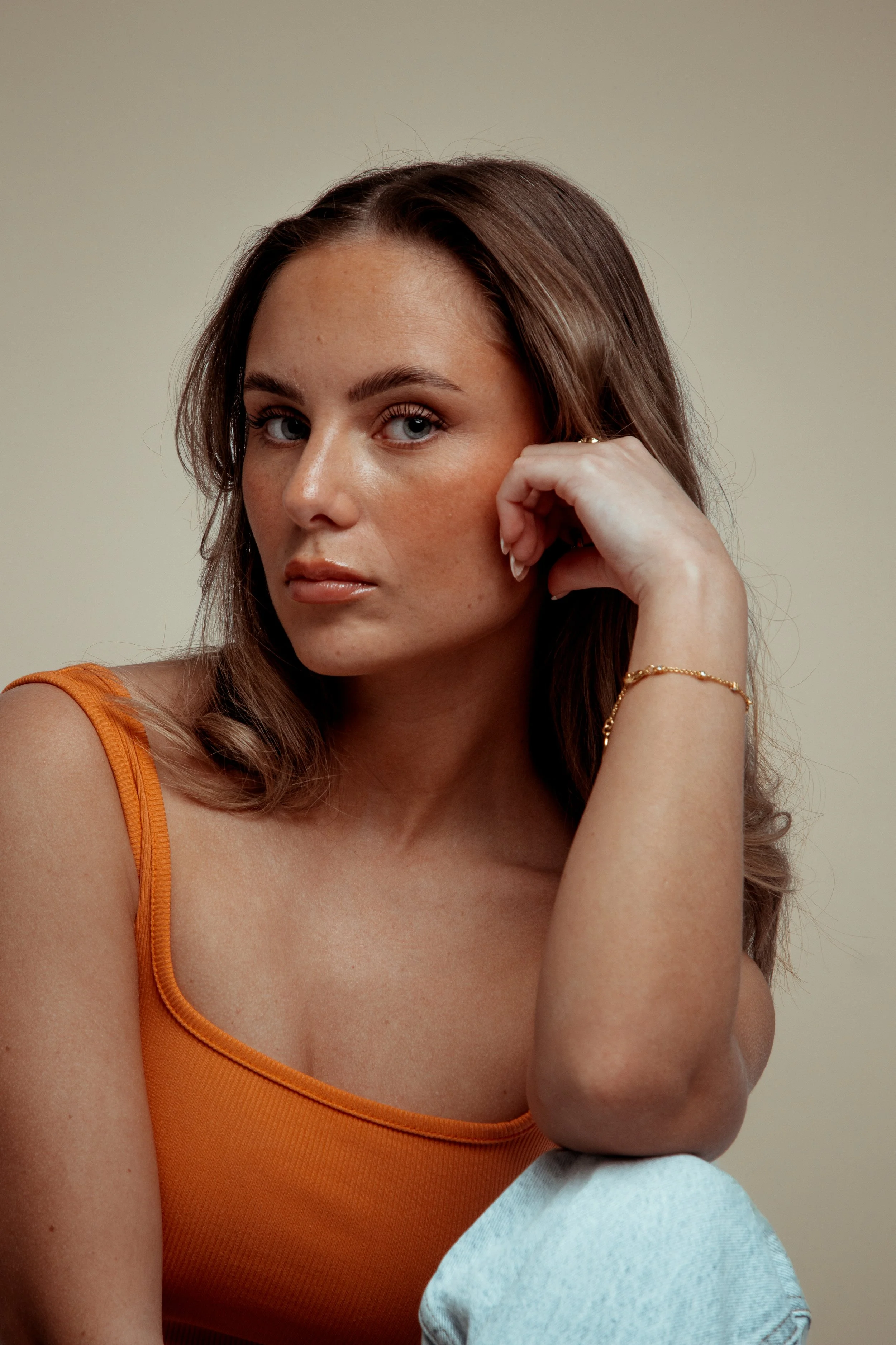 Portrait of a woman with long brown hair, wearing an orange tank top, resting her head on her hand, against a plain beige background.