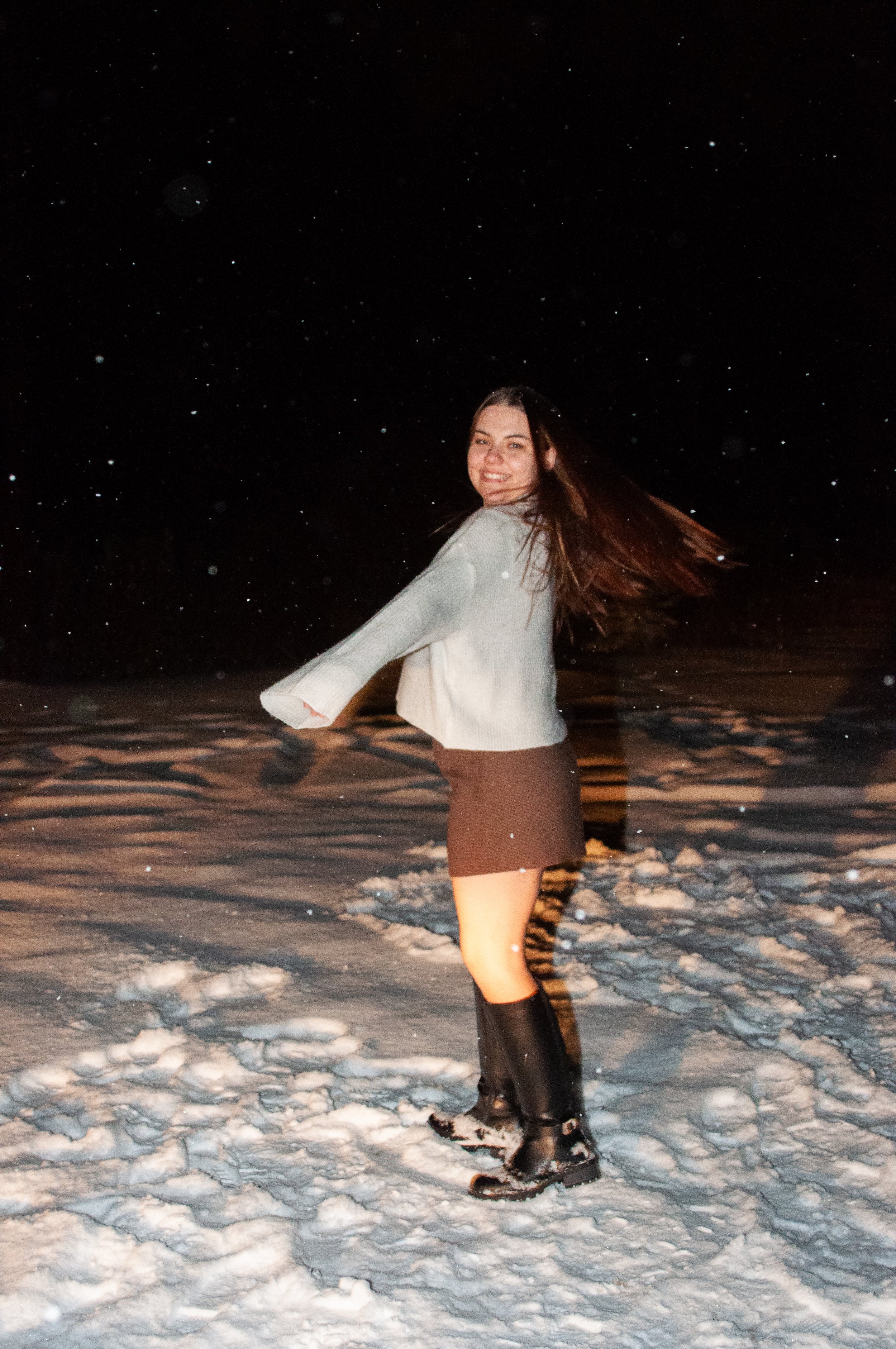 A woman smiling and spinning in the snow at night, wearing a skirt, a light-colored sweater, and knee-high snow boots.