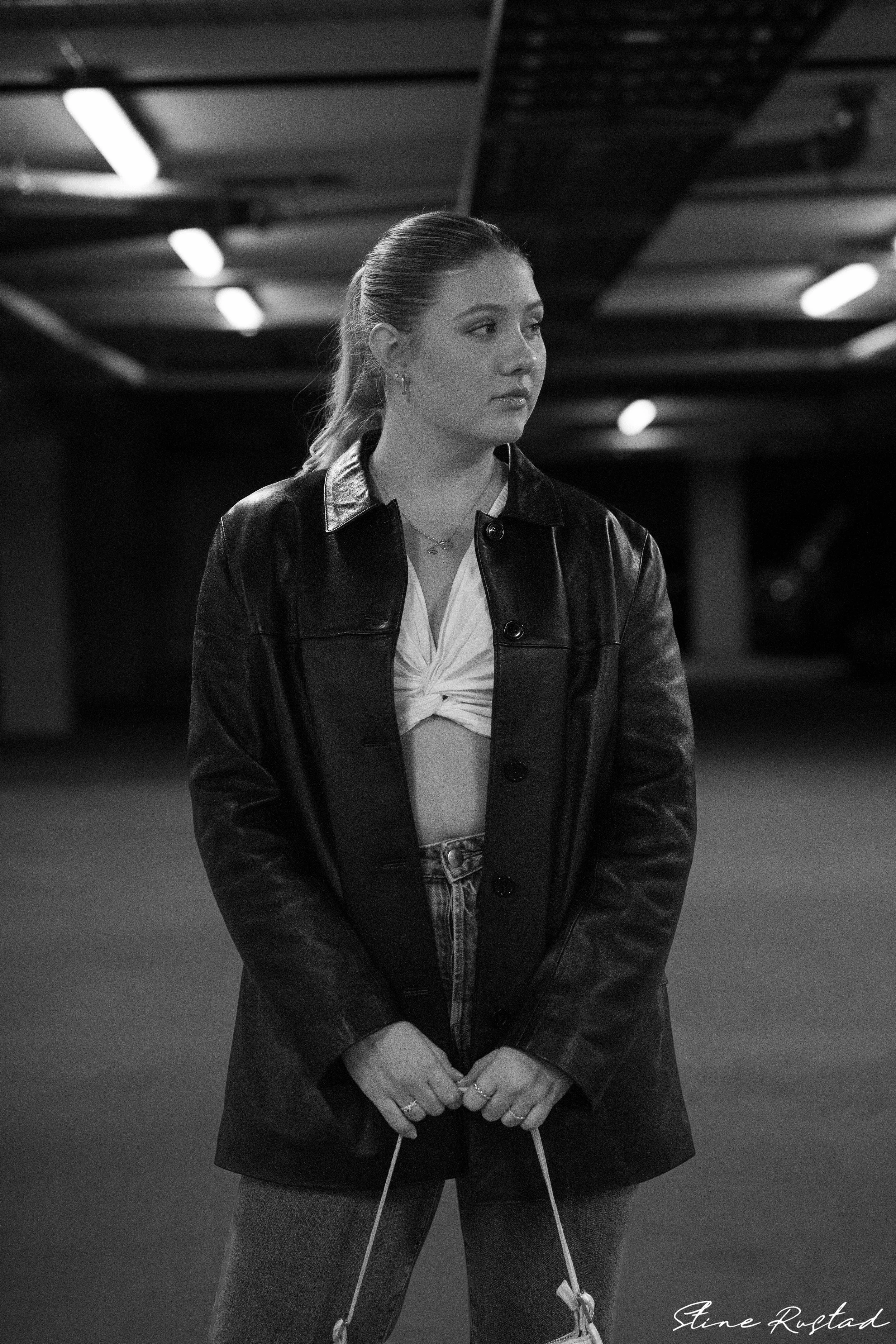 A young woman standing in an indoor parking garage, wearing a leather jacket, a knotted white top, and jeans, looking to her right.