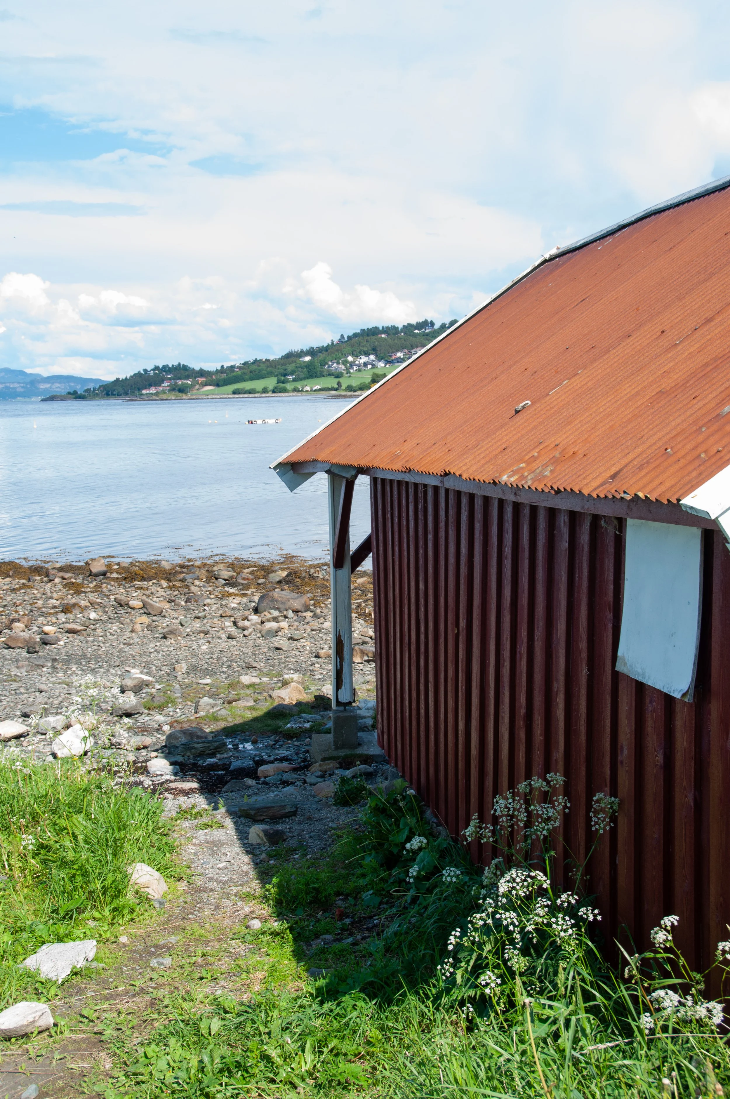A small wooden building with a rusty metal roof on a rocky shoreline, with water and hilly landscape in the background under a partly cloudy sky.