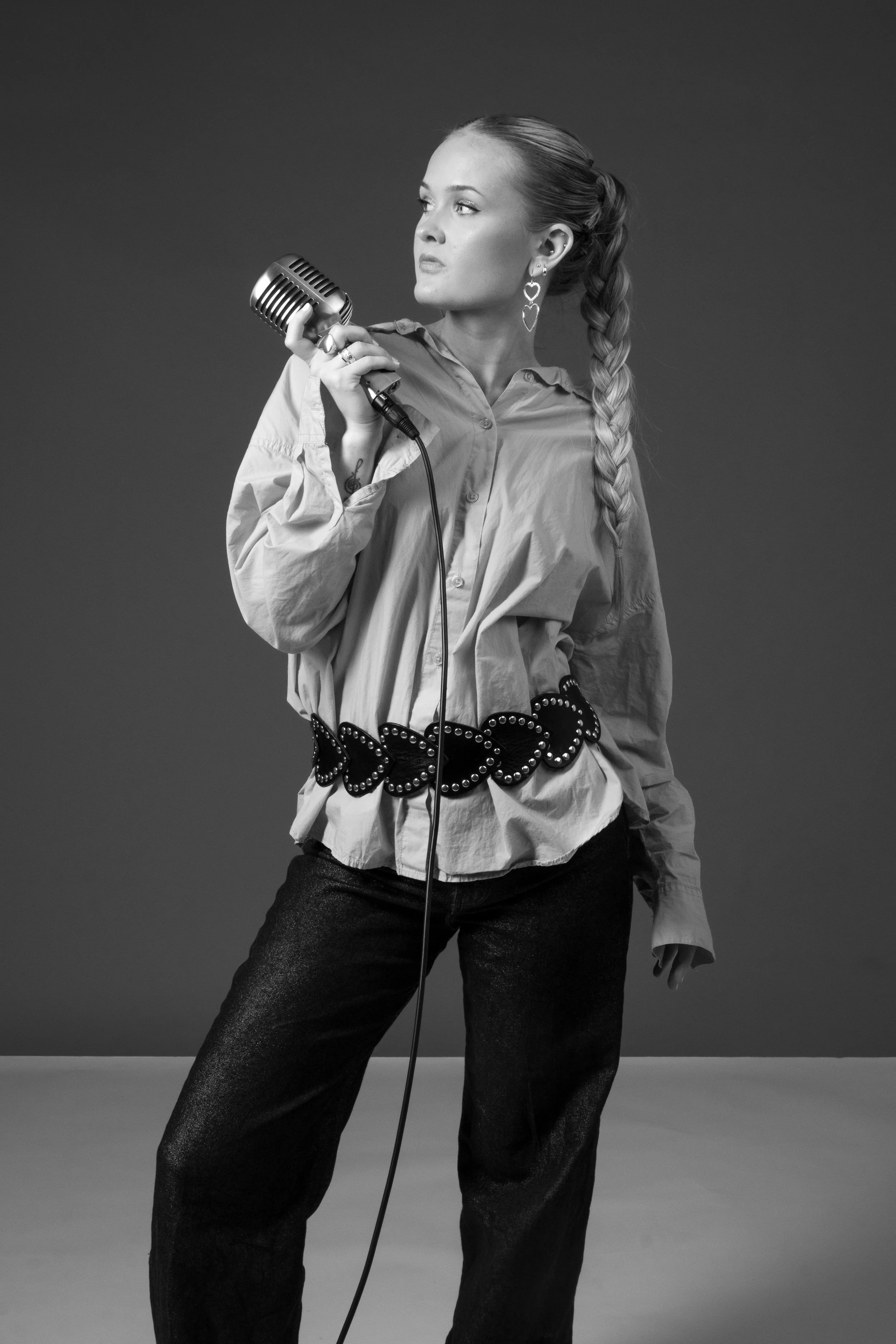 Black and white photo of a woman with long braid holding a vintage microphone, dressed in a loose button-up shirt and dark pants, standing against a plain background.