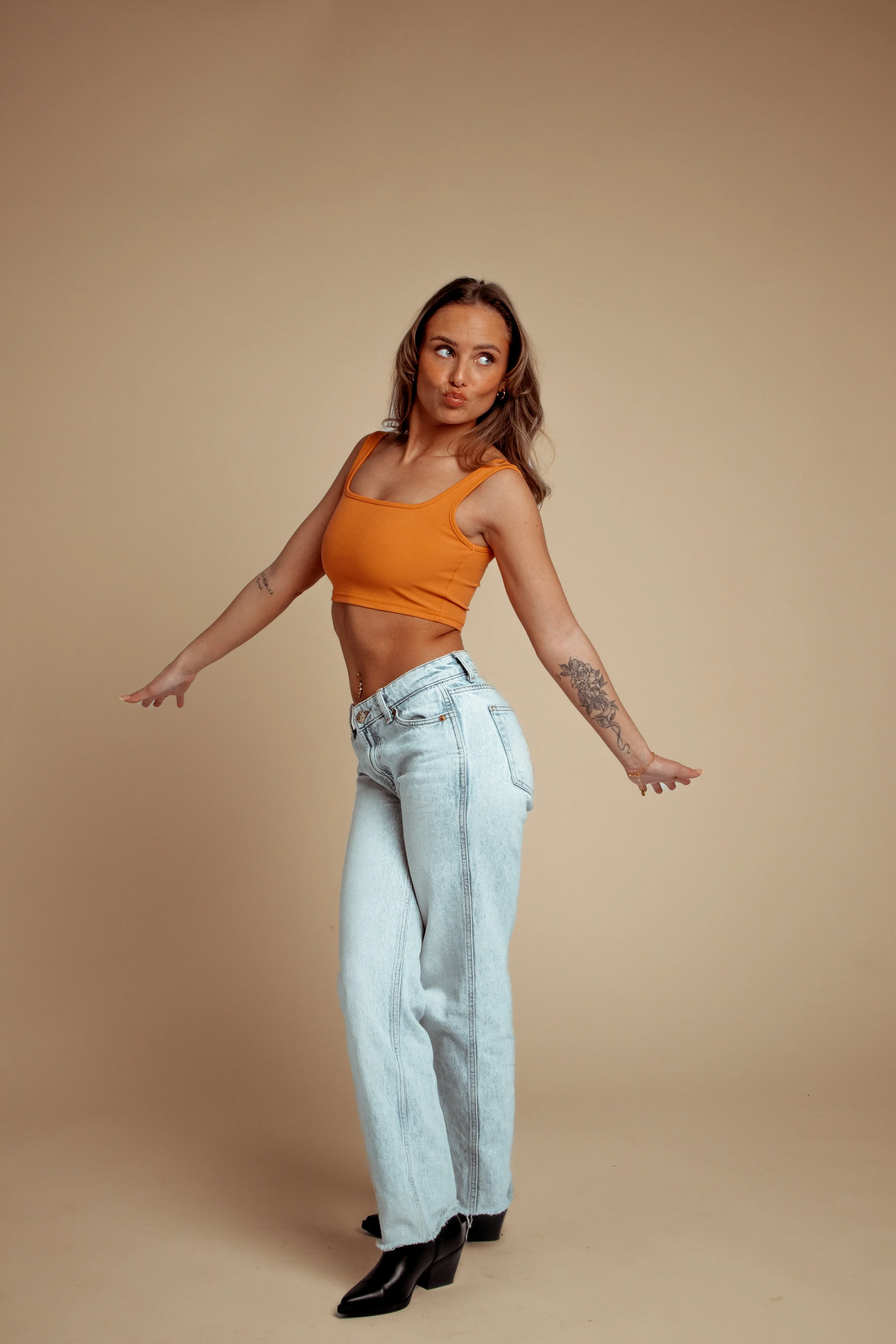 A young woman with light brown hair in loose waves wearing an orange crop top, high-waisted light blue jeans, and black boots posing with her arms extended to the sides against a plain beige background.