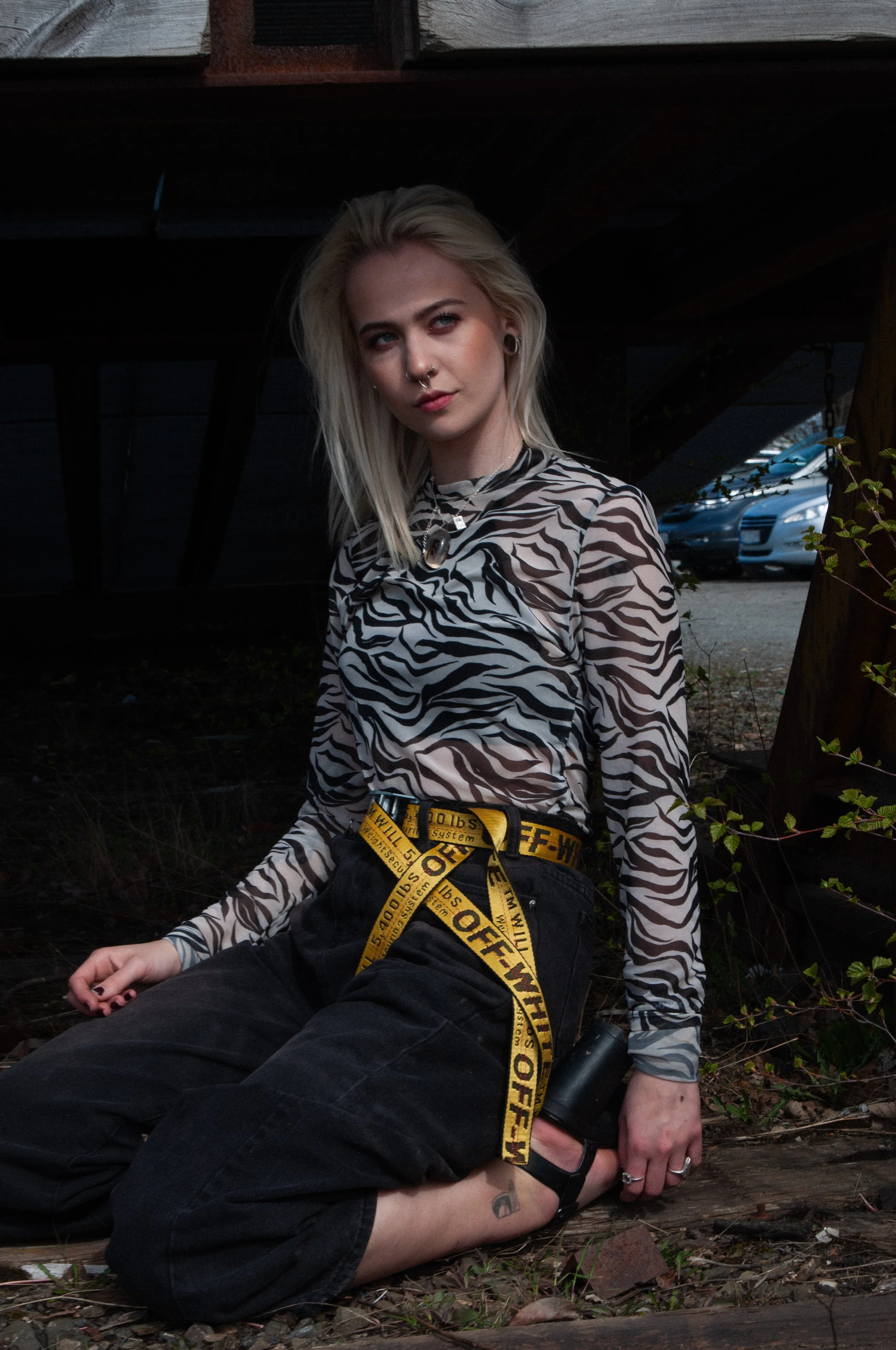 Young woman with platinum blonde hair, dark makeup, and multiple face piercings, wearing a zebra-print sheer top, black cargo shorts, and a yellow Off-White belt, sitting on the ground near a wooden structure, with a parking lot and cars in the backg