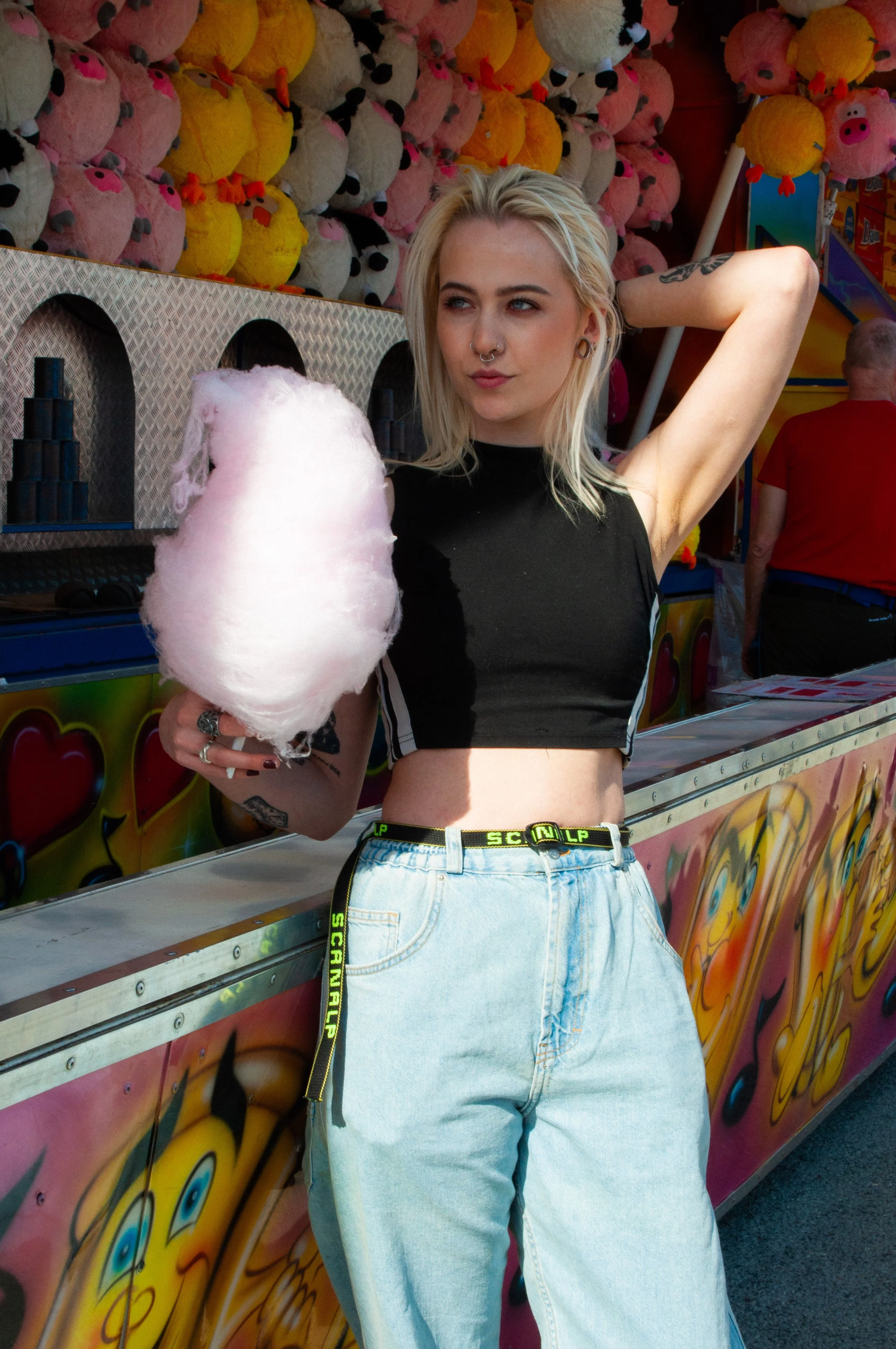 A young woman with blonde hair, black crop top, and high-waisted light blue jeans holding pink cotton candy at a carnival game booth with stuffed animal prizes in the background.