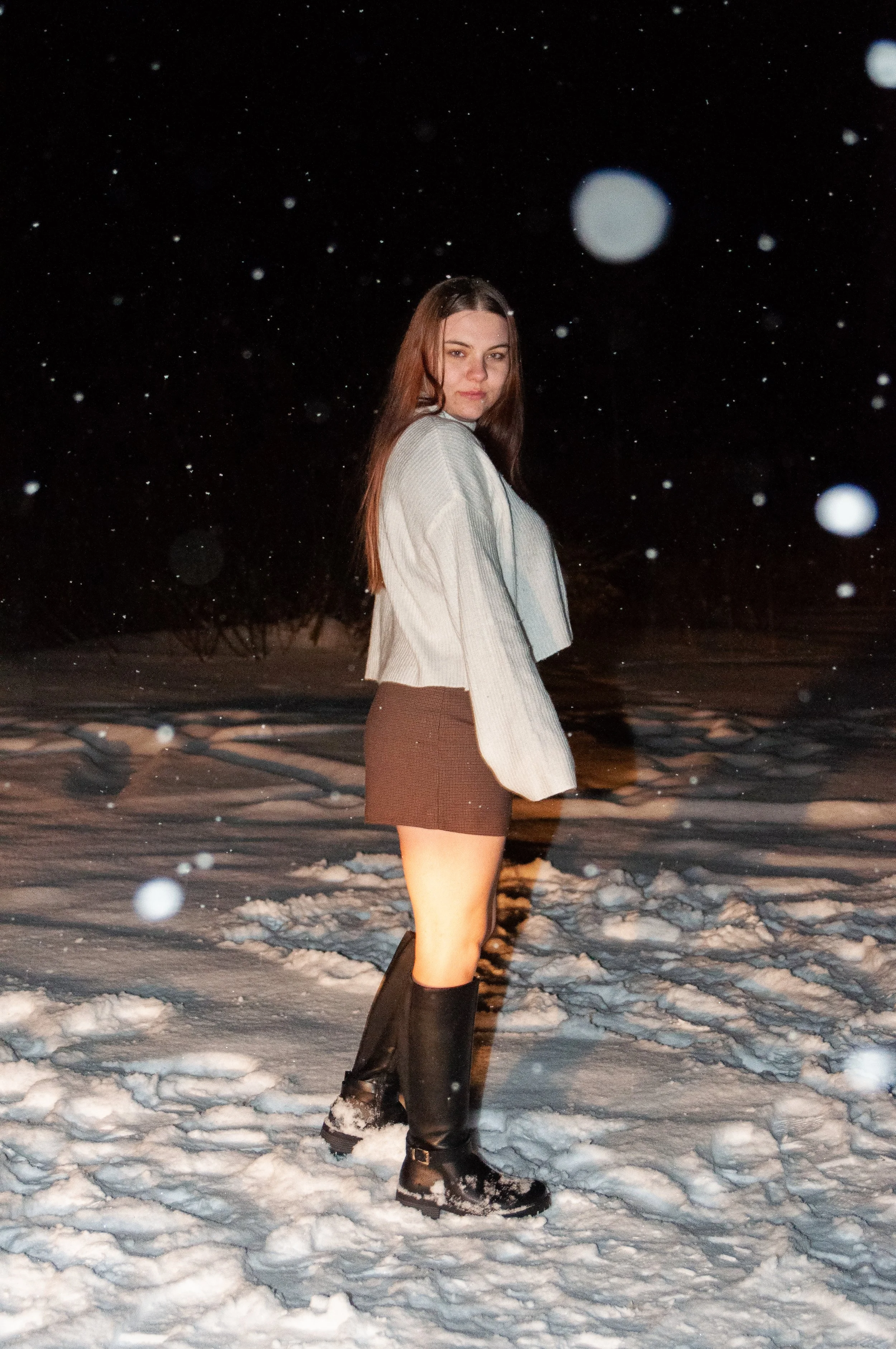 A young woman standing in a snow-covered outdoor area at night, wearing a gray sweater, a dark skirt, and tall black boots, with snow falling around her.