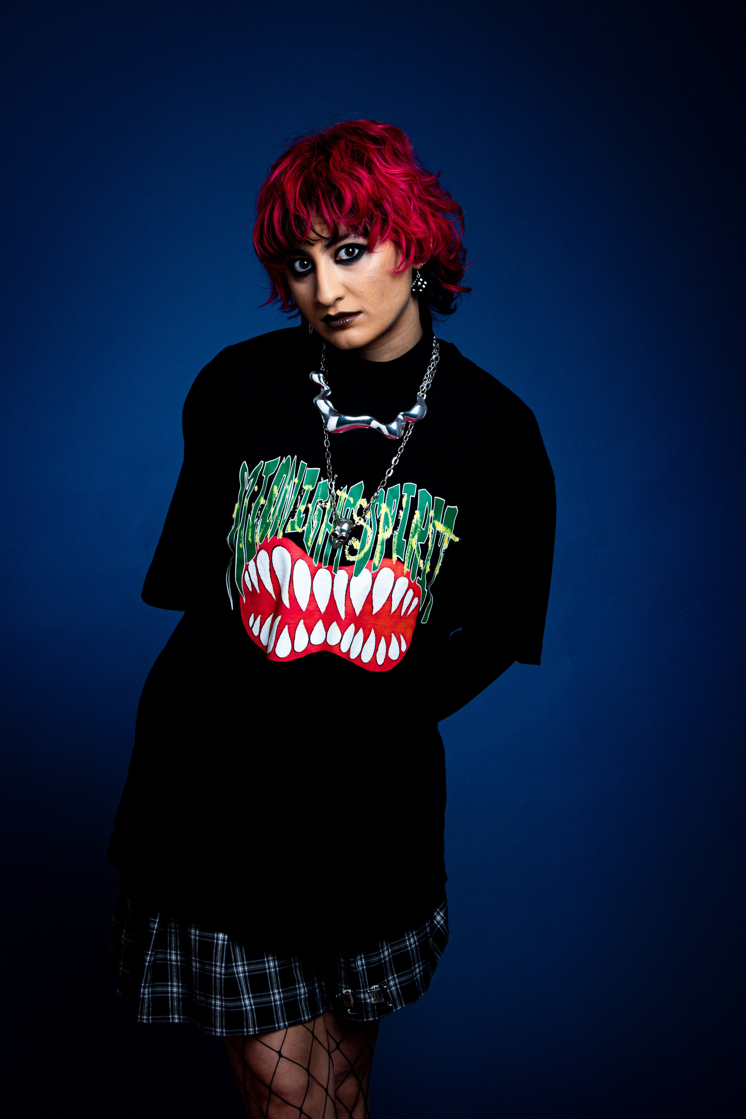 A young woman with short, vibrant pink hair looking at the camera, wearing dark lipstick, a black graphic t-shirt with a colorful monster mouth design, a plaid skirt, fishnet stockings, and bold jewelry, against a dark blue background.
