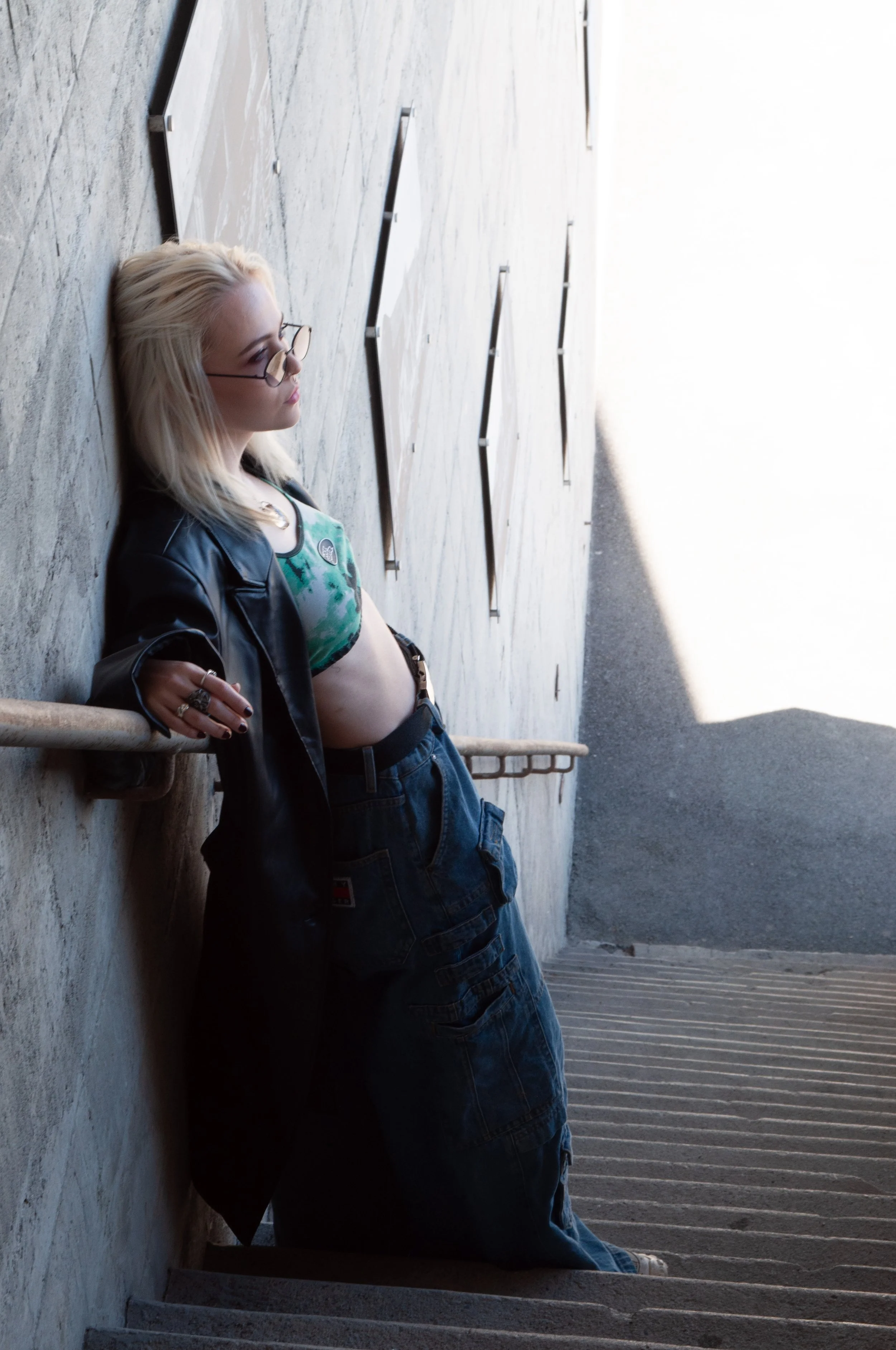 A young woman with platinum blonde hair and glasses leaning against a concrete wall on a stairway, wearing a black jacket, a green tie-dye crop top, and baggy blue pants.
