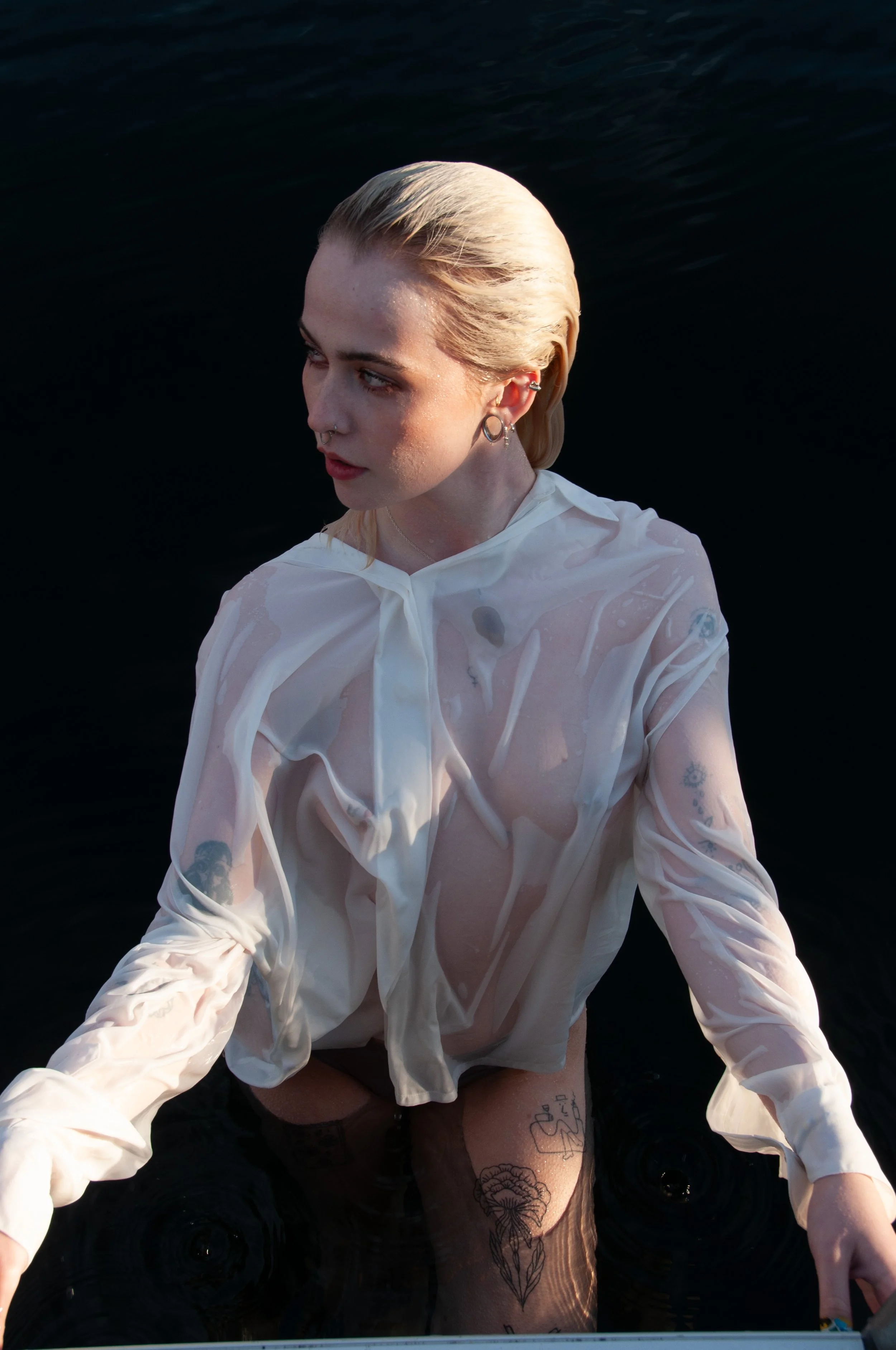 A woman with platinum blonde hair and tattoos submerged in water, wearing a white, wet, semi-transparent shirt.