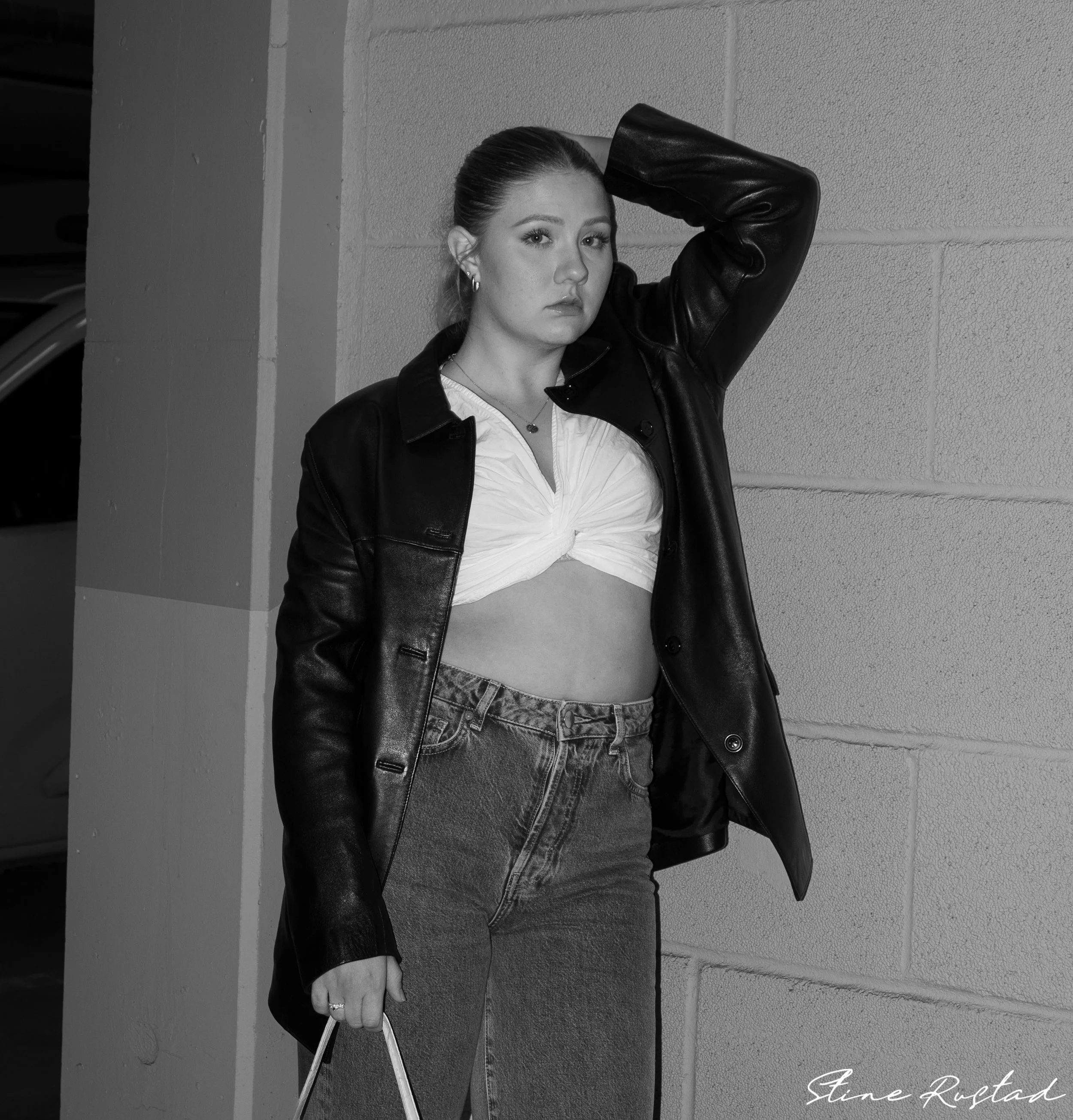 A young woman with slicked-back hair in a black leather jacket, white cropped top, and high-waisted jeans, standing against a wall and posing with one hand behind her head.