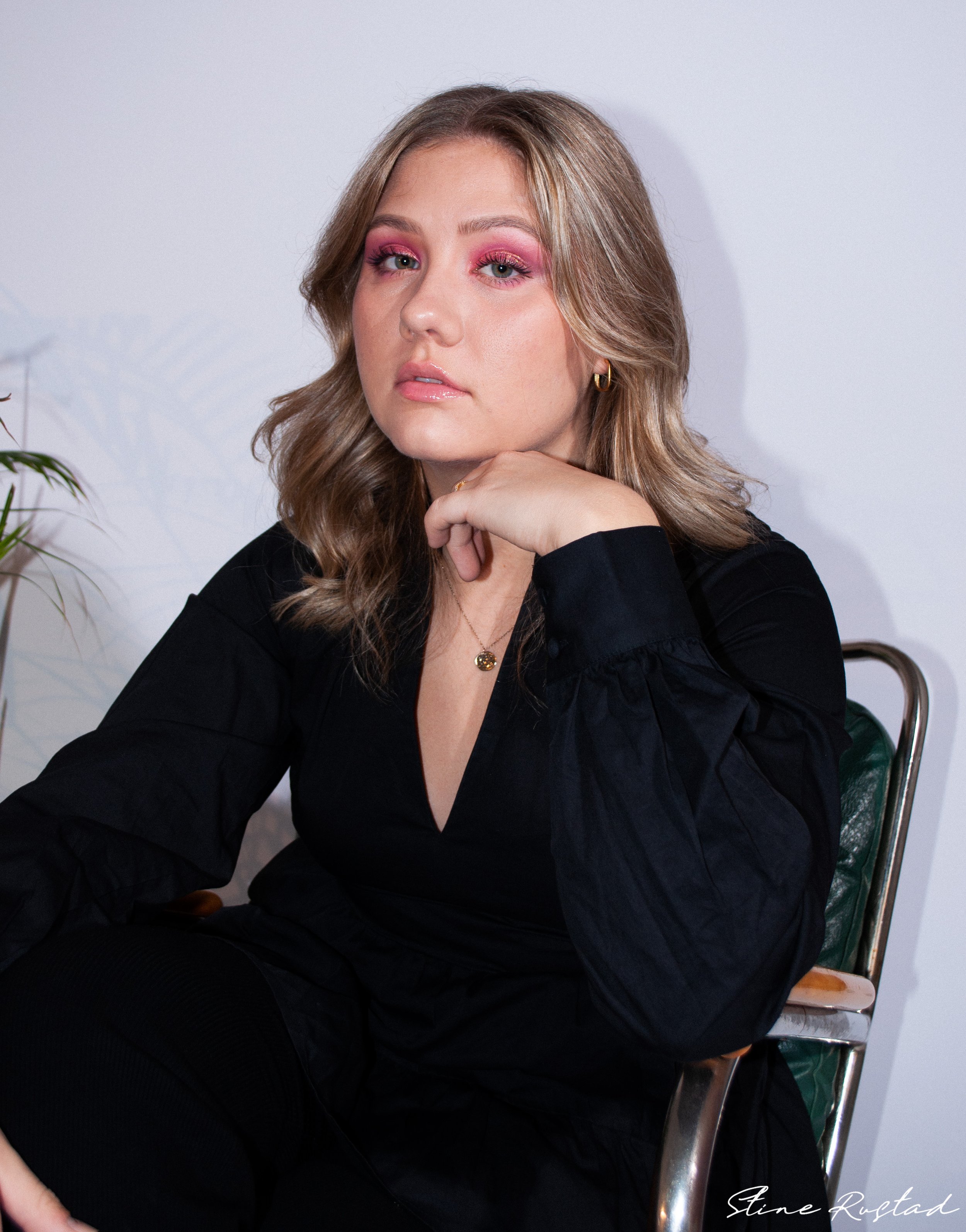 Young woman with blonde hair, pink eyeshadow, gold hoop earrings, and a gold necklace, sitting on a chair against a plain white wall.