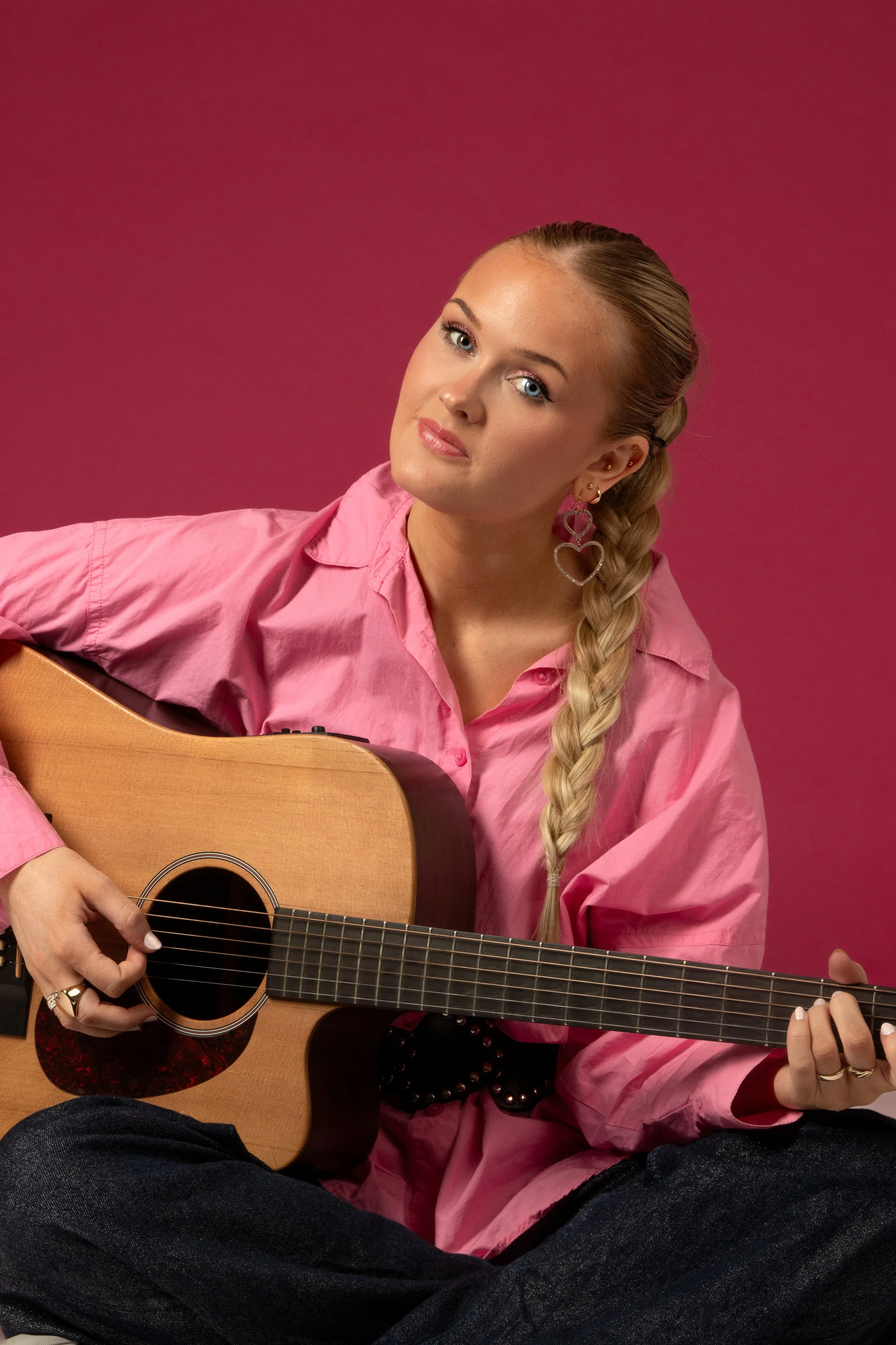 A woman with blonde hair in a braid, wearing a pink shirt, playing an acoustic guitar against a pink background.