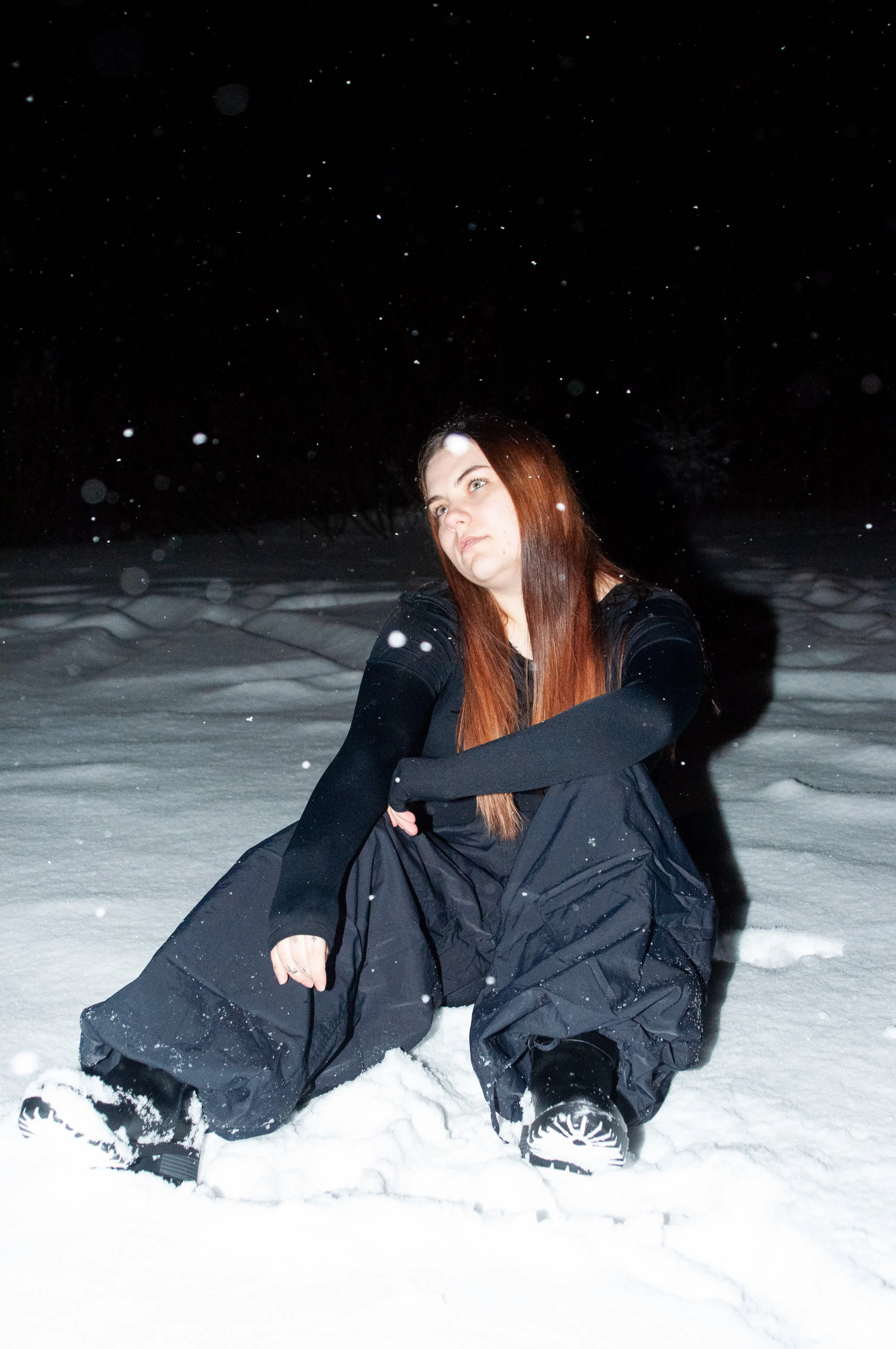 A woman with long red hair sitting in deep snow at night, dressed in black outdoor clothing, with snow falling around her.