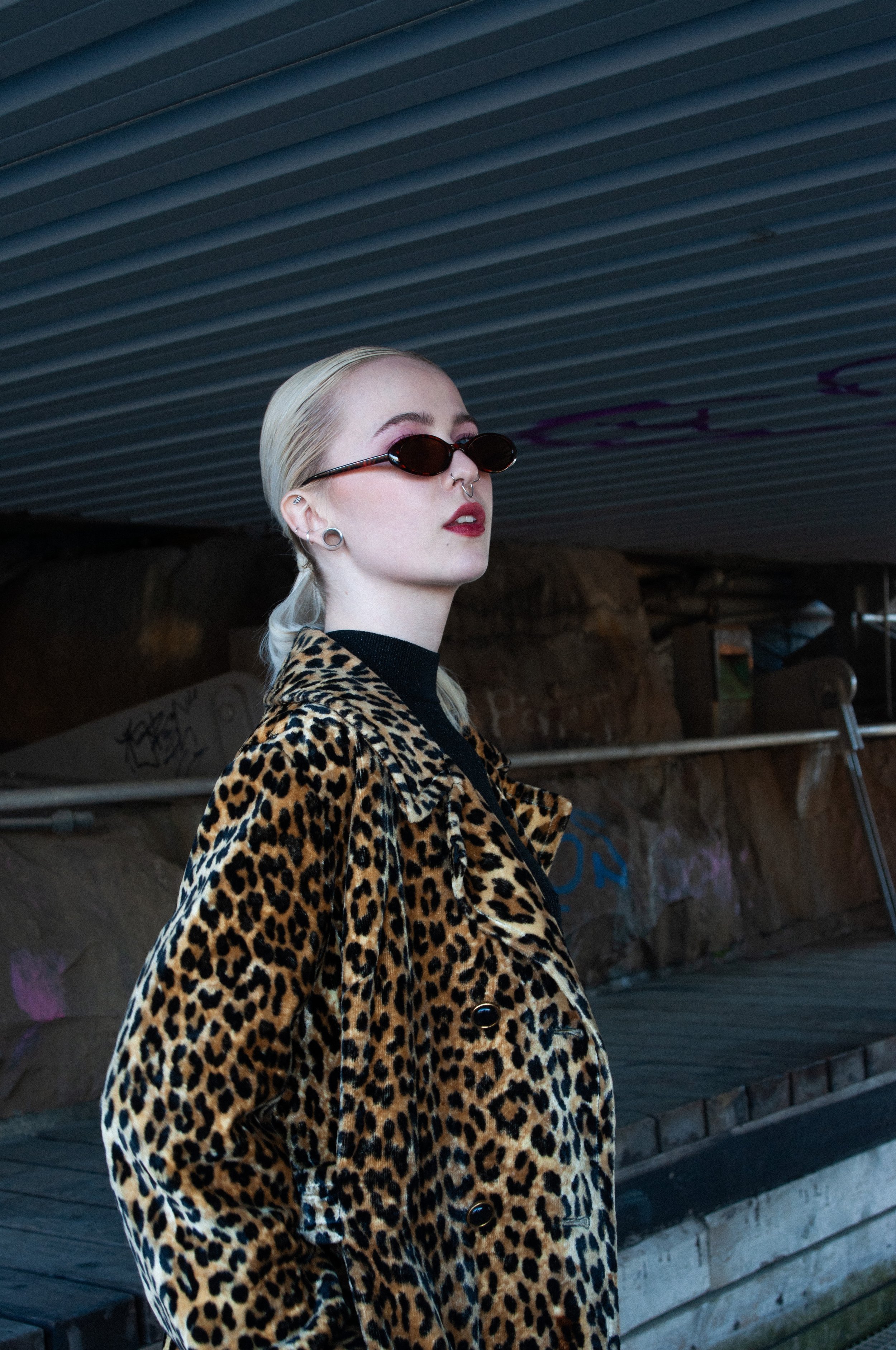 A woman with platinum blonde hair, wearing black oval sunglasses, a leopard print coat, and dark lipstick, standing under a metallic overhead structure.